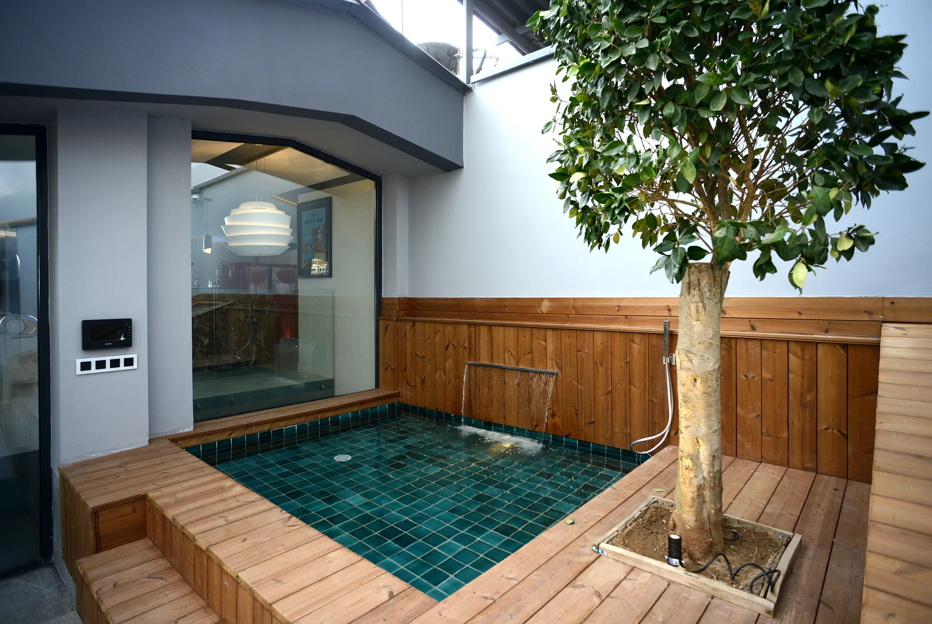 A small square plunge pool with teal tiles is sunken into a wooden deck alongside a tall green tree and a waterfall spout.