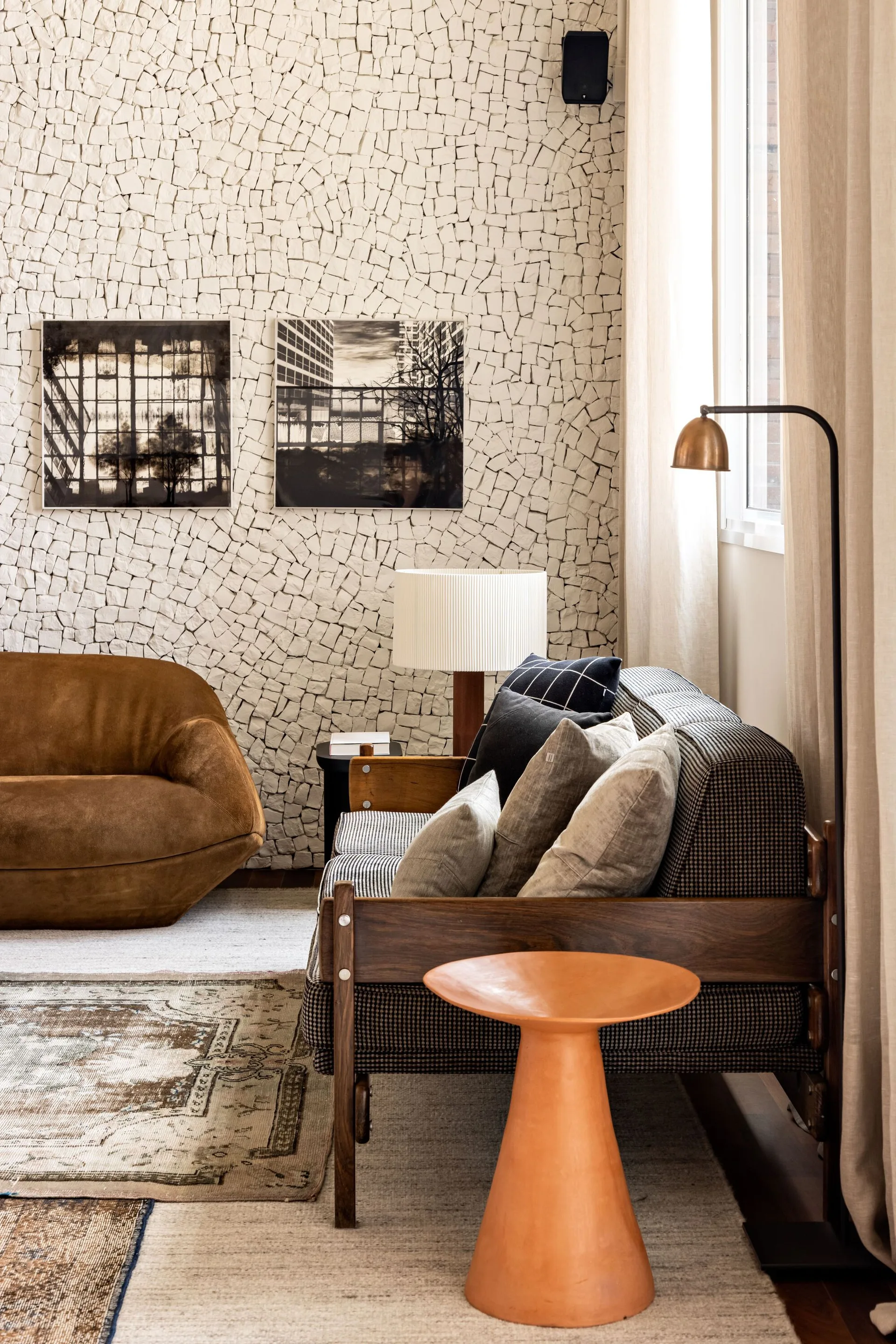 A living room features a wooden frame sofa, a rounded brown armchair, and a terracotta side table against a textured stone mosaic wall.