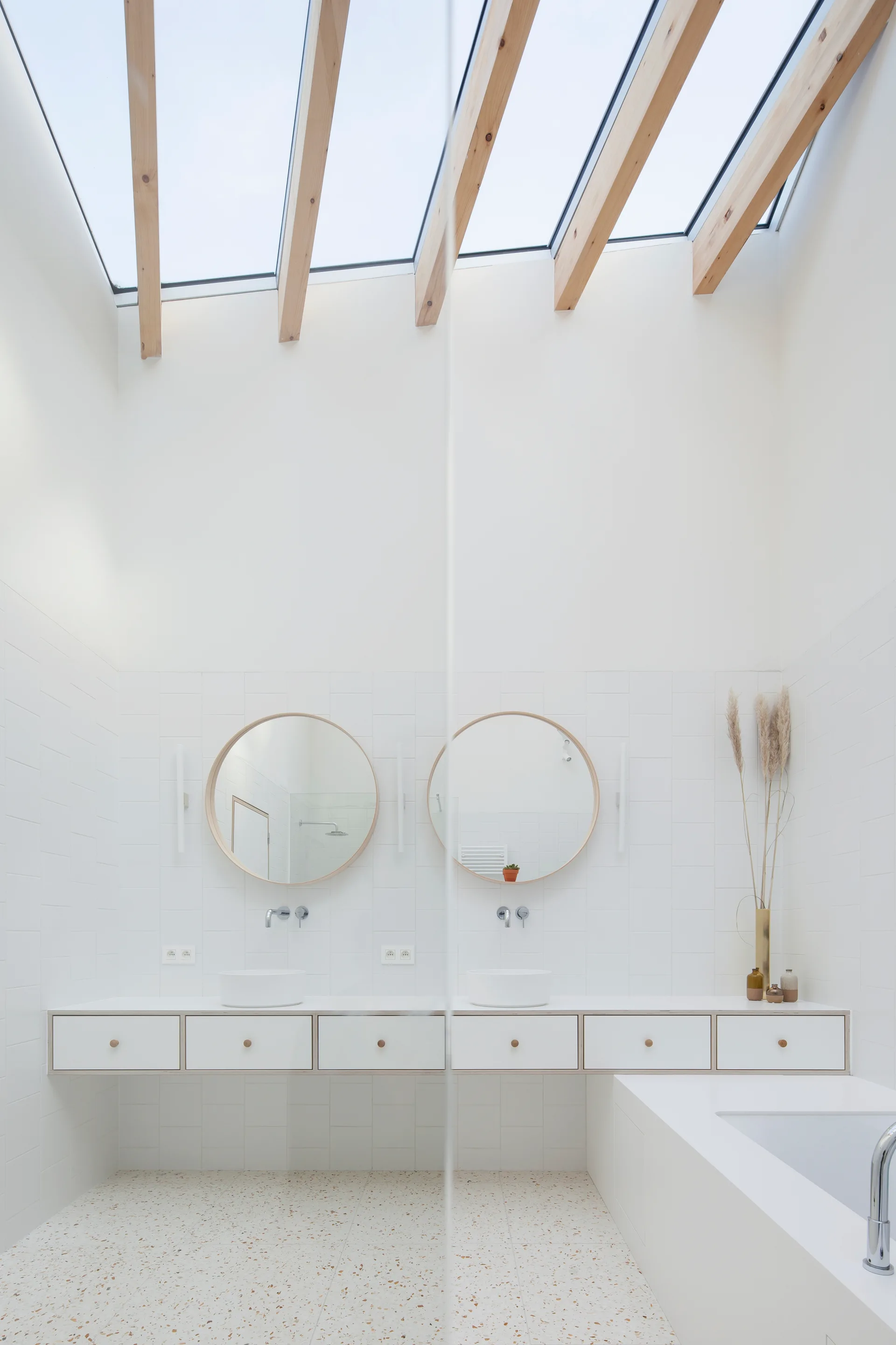A minimalist white bathroom features a floating double vanity, circular wood-framed mirrors, and a large ceiling skylight with exposed wooden rafters.