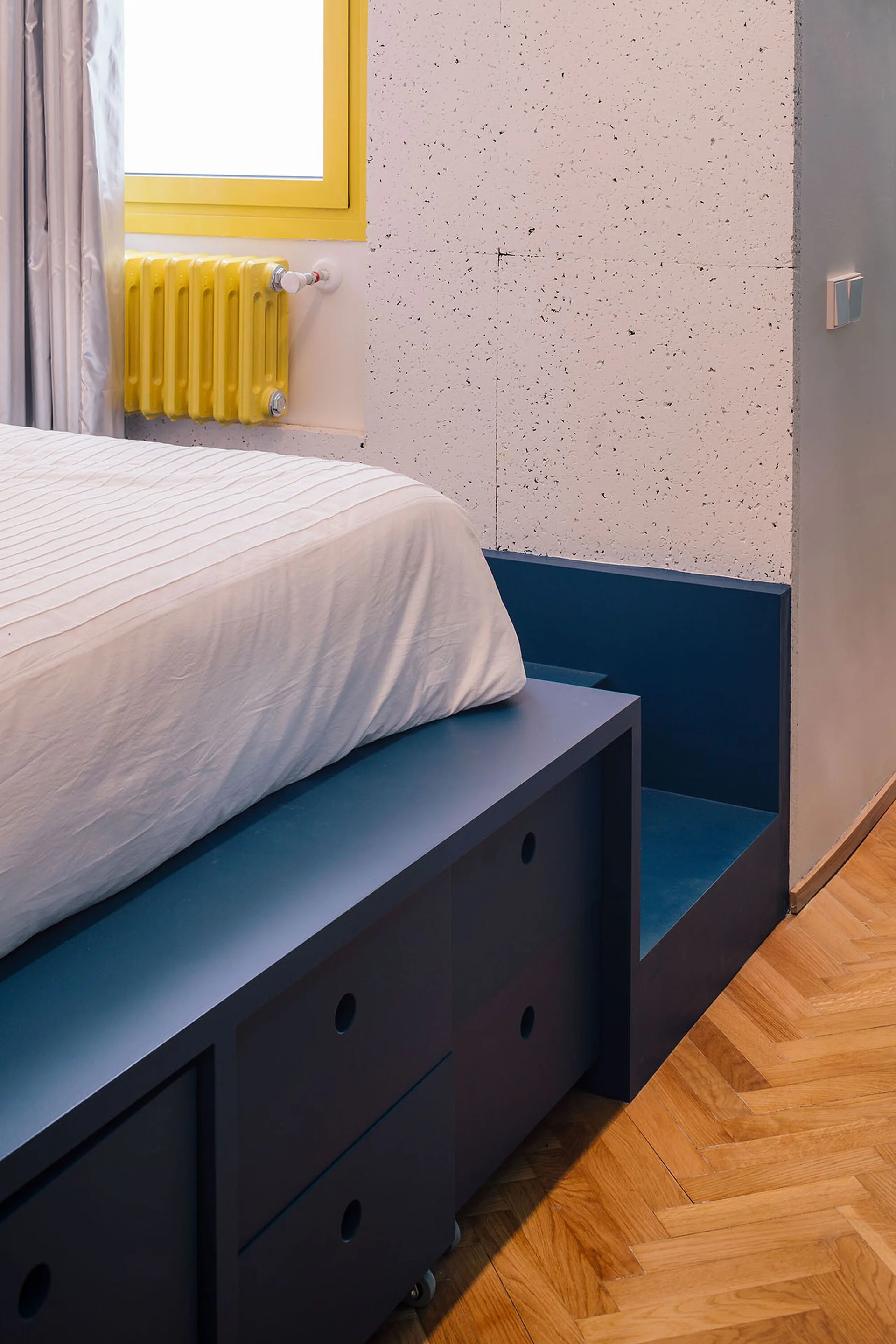 A blue built-in platform bed with storage drawers sits on a herringbone wood floor next to a bright yellow radiator and window.