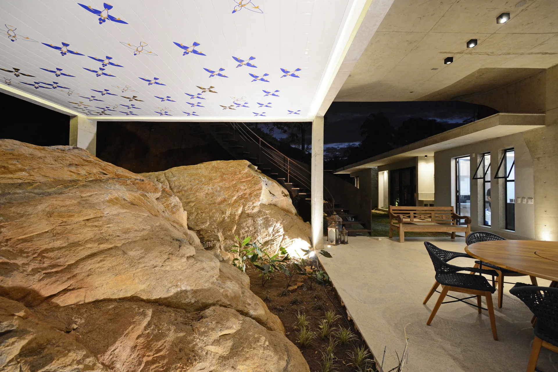 A covered concrete patio at night features a large illuminated natural rock formation next to an outdoor dining table and chairs.