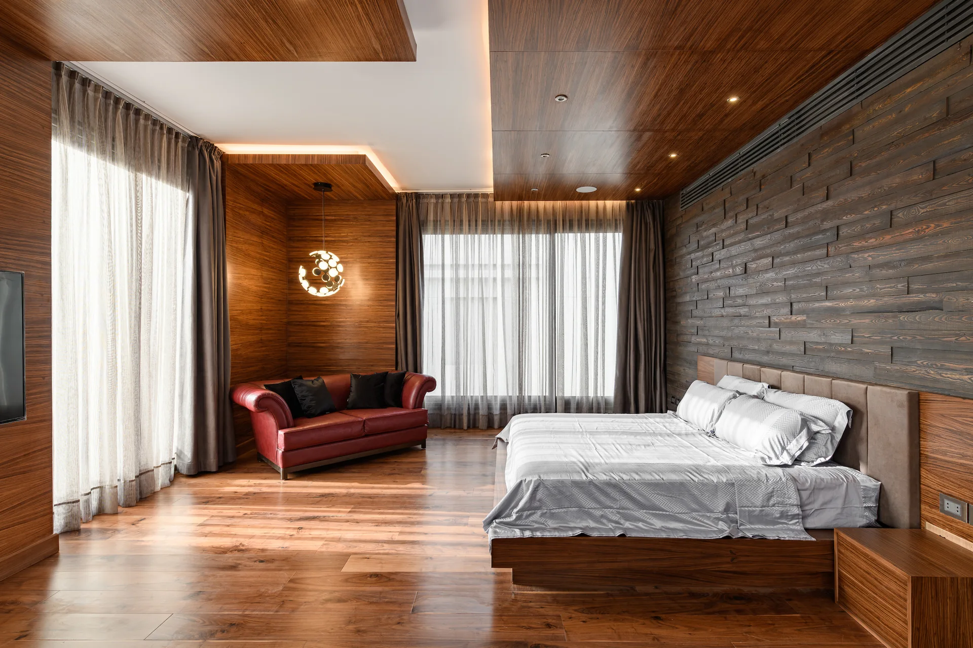 A modern bedroom features extensive wood paneling on the ceiling, a dark wood slat accent wall, a platform bed, and a red leather sofa in the corner.