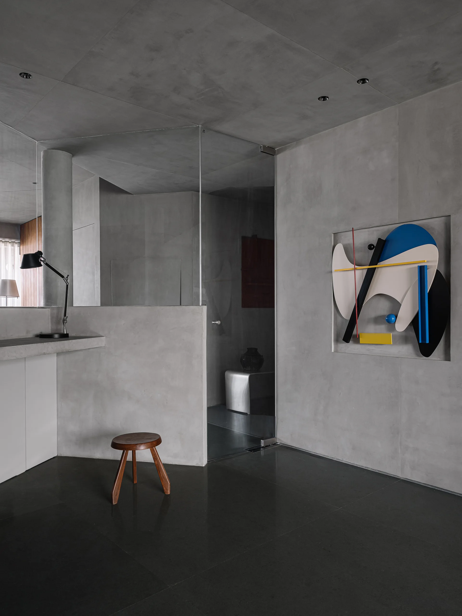 A minimalist concrete interior features a recessed geometric wall sculpture, a dark wood stool, and a built-in concrete desk.