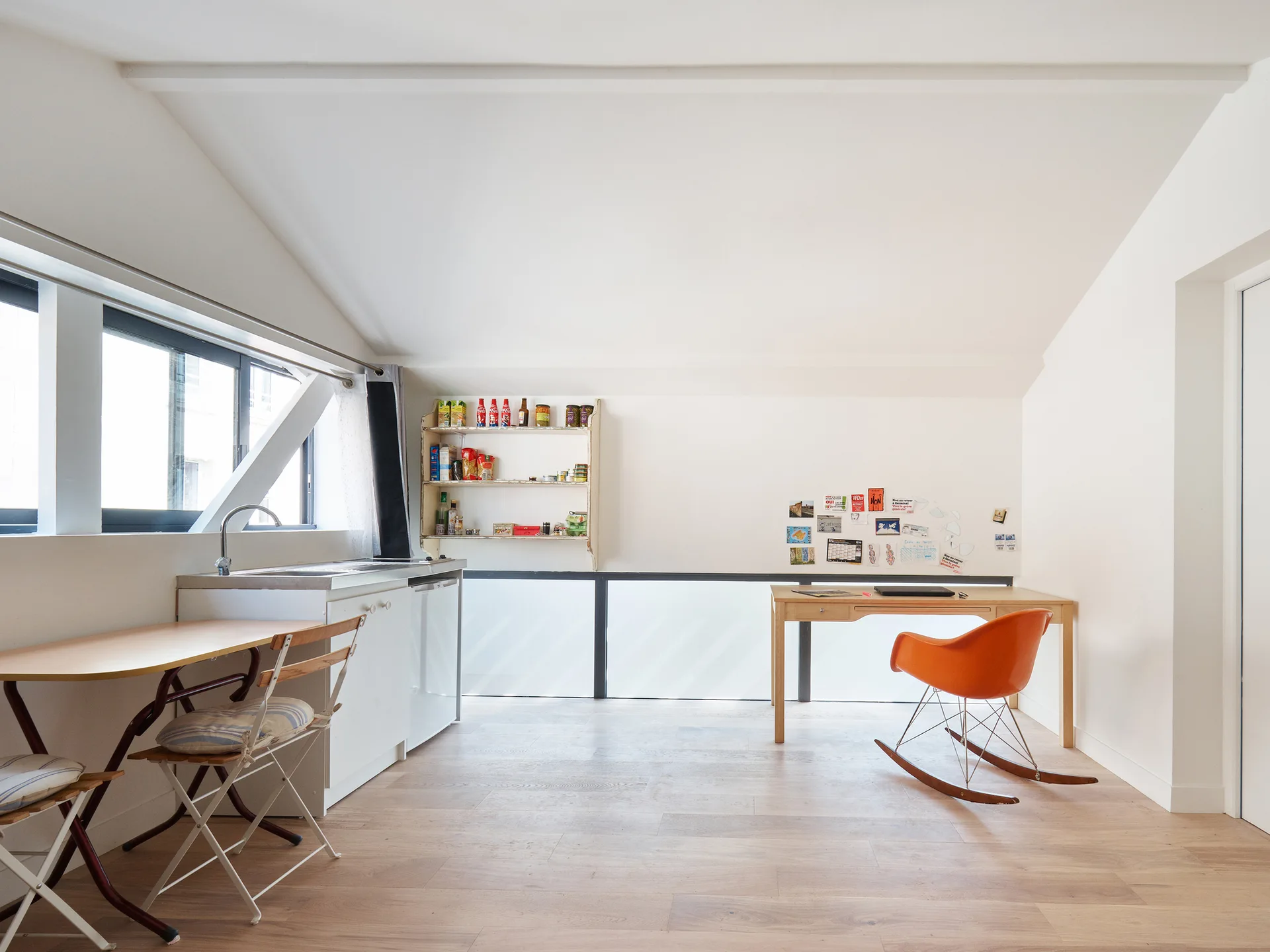 A bright attic studio features a white kitchenette on the left and a wood desk with an orange rocking chair on the right under a sloped ceiling.
