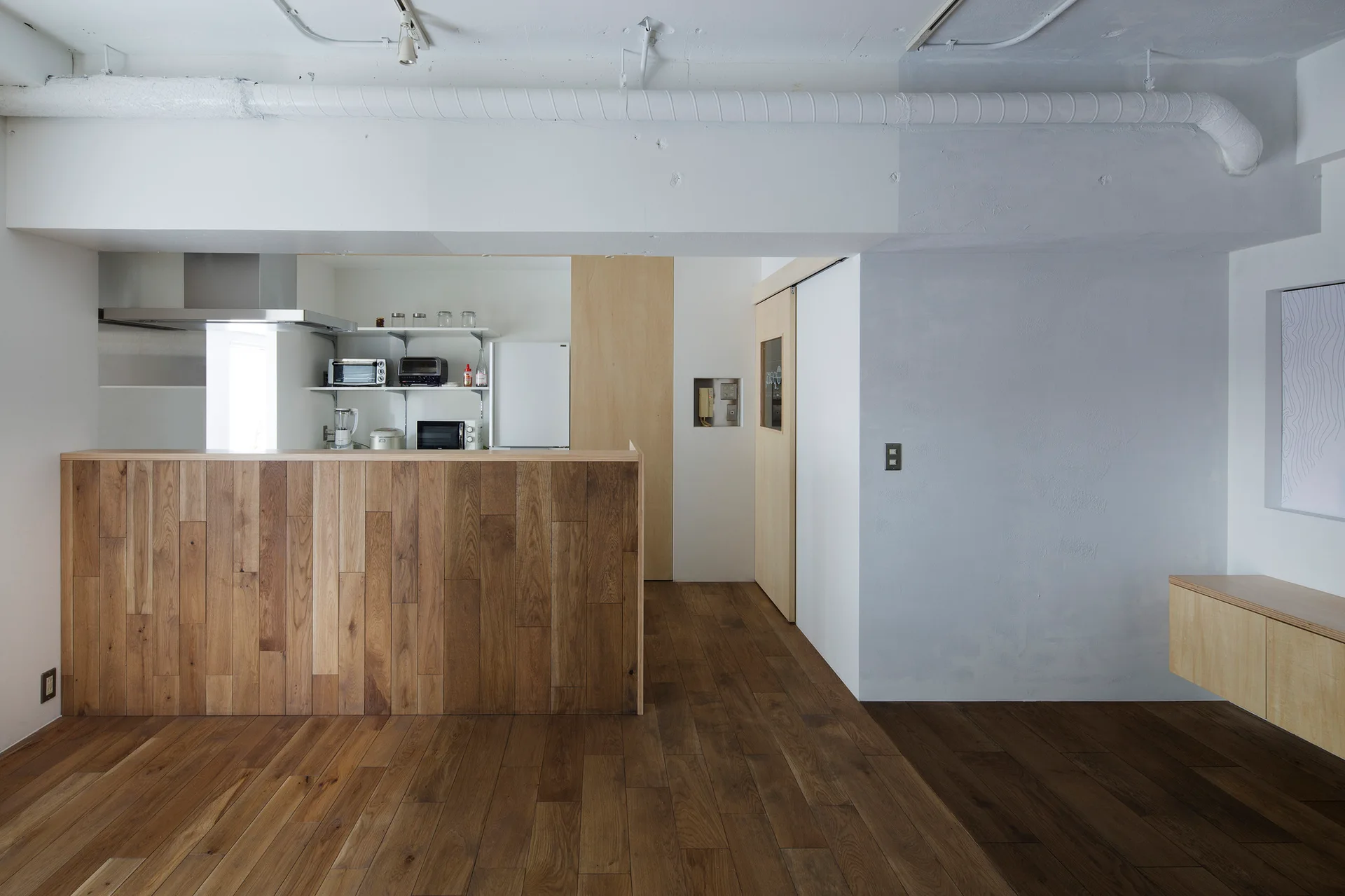 A minimalist kitchen features a wood-paneled counter, matching hardwood floors, a sliding wood door, and a white ceiling with exposed painted pipes.