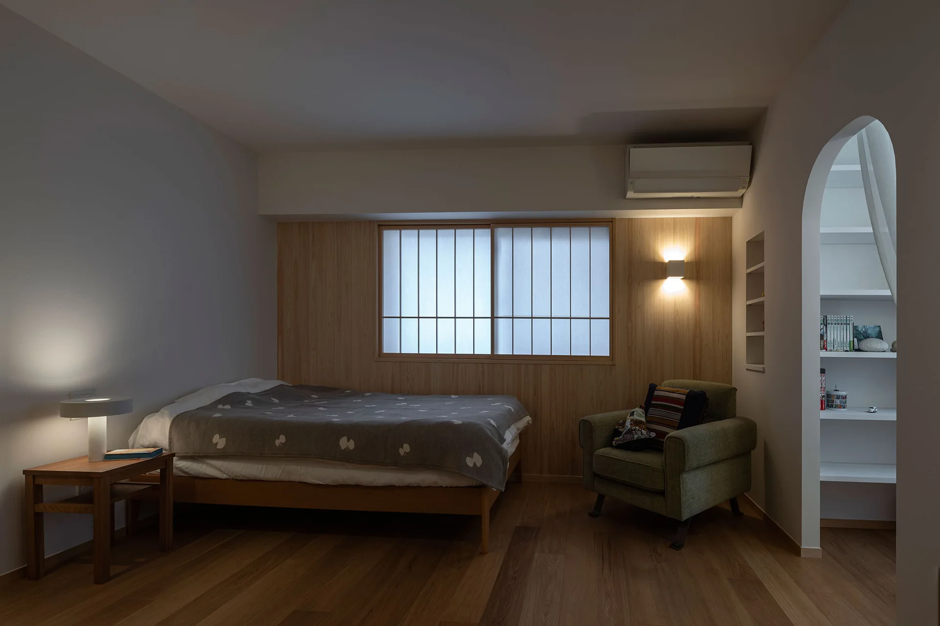 A minimalist bedroom features a low wooden bed, a green armchair, a shoji-inspired window on a wood-paneled wall, and an arched entryway.