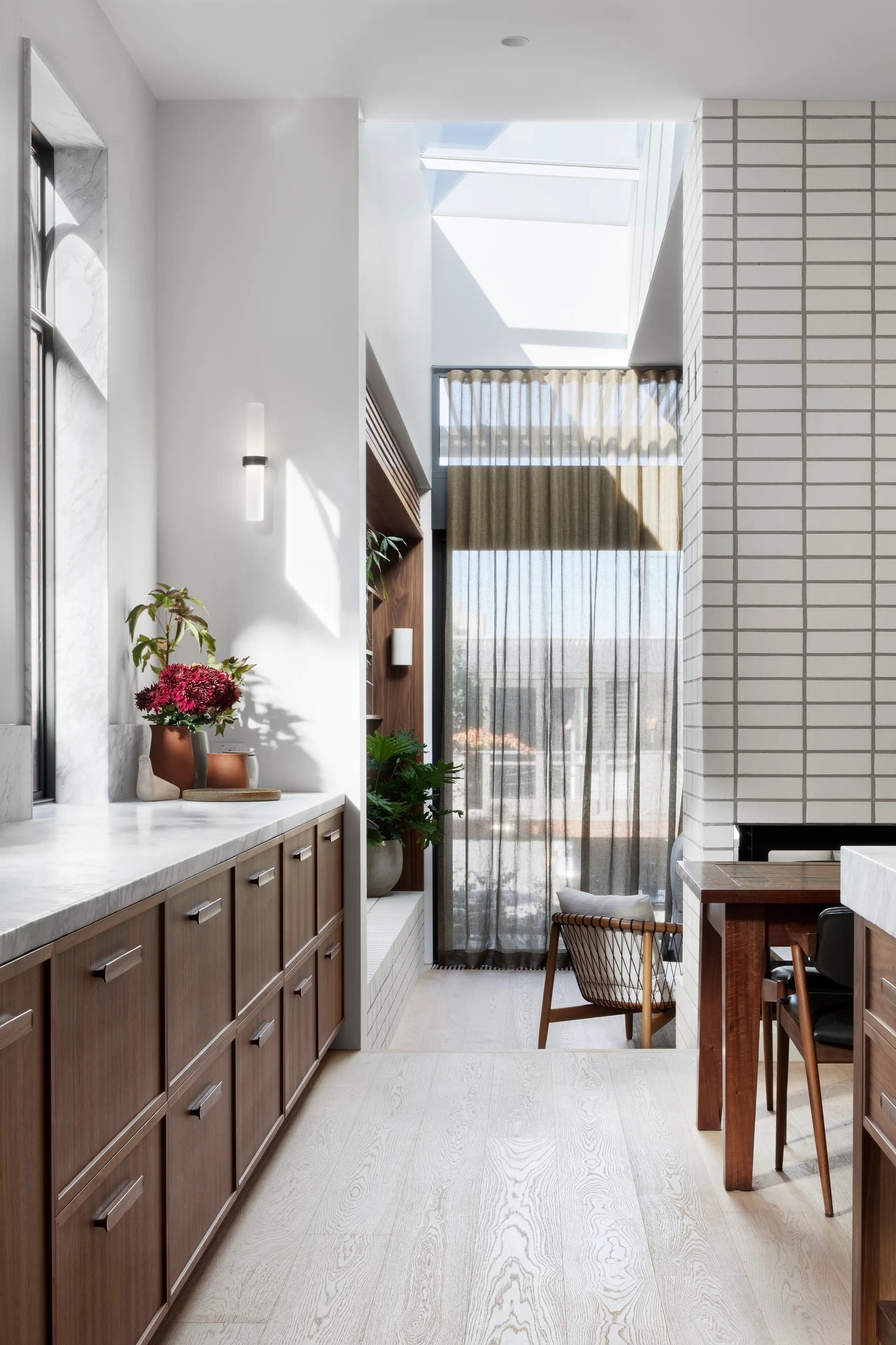 A modern kitchen features dark wood cabinetry with a marble countertop, adjacent to a dining area with a white grid-tiled pillar and a skylight.