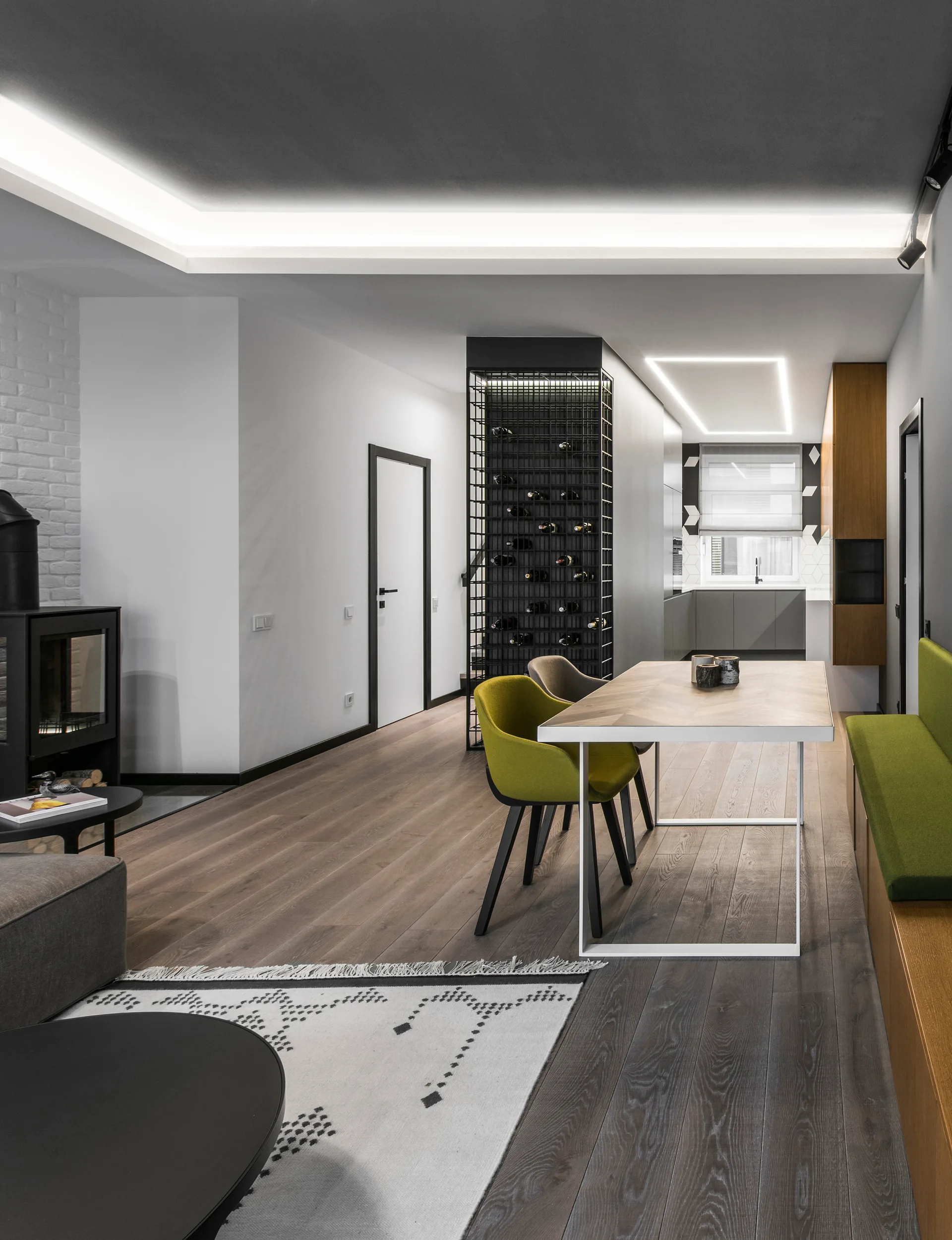 A modern dining room features a herringbone wood table, chartreuse chairs, a green upholstered bench, and a tall black metal wine storage tower.