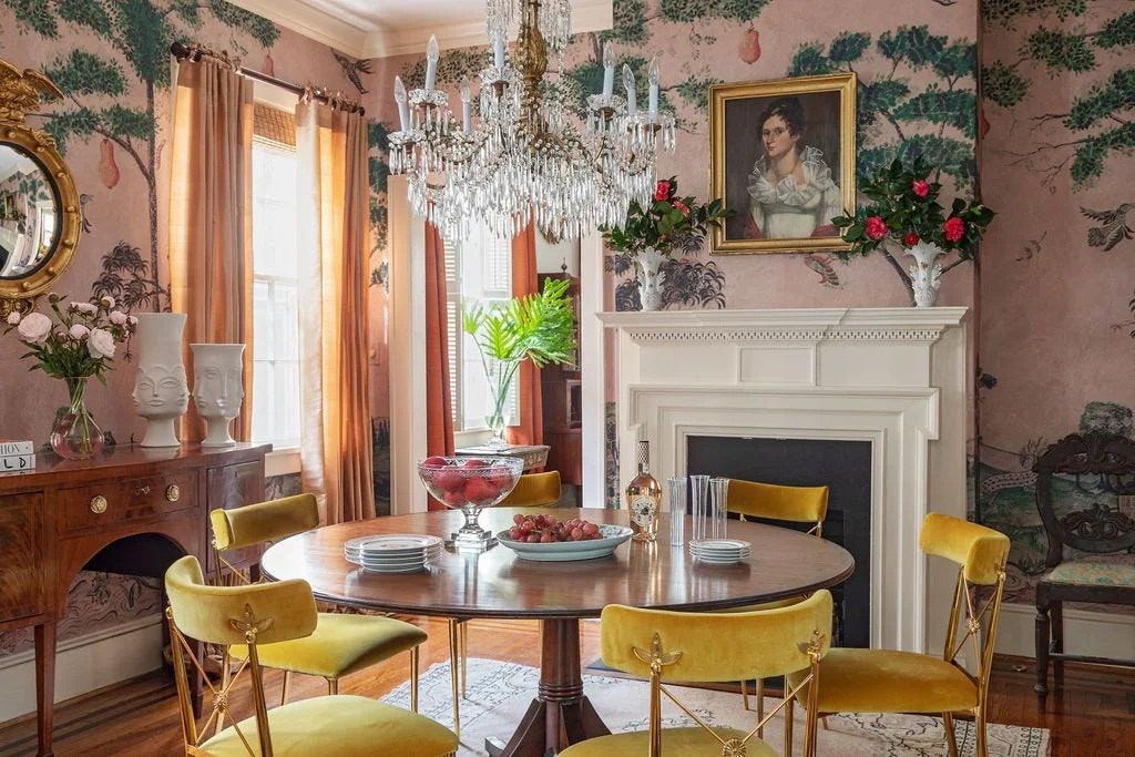A formal dining room features pink landscape wallpaper, yellow velvet chairs around a round wood table, and an ornate crystal chandelier.