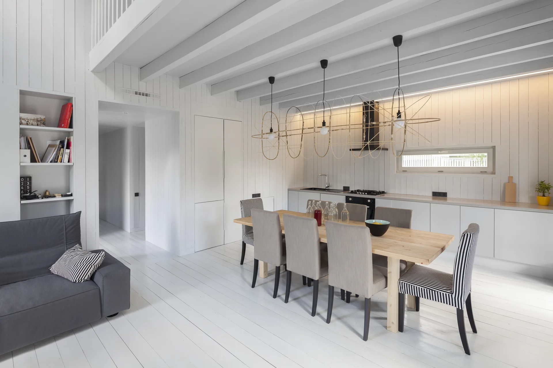 A white paneled kitchen and dining space featuring a light wood table, grey and striped chairs, and sculptural wire pendant lights.