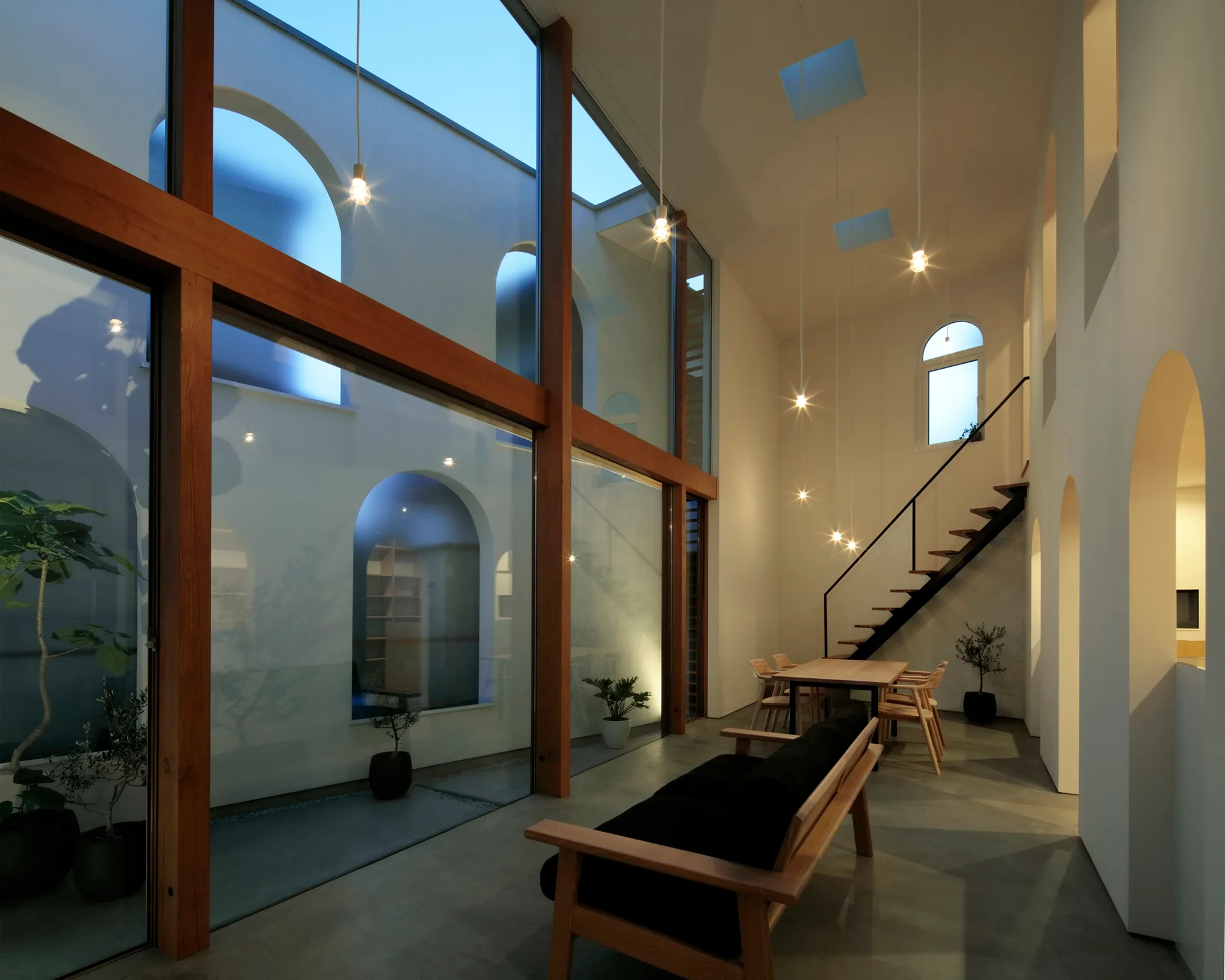 A minimalist double-height living room features large wood-framed windows, an open-riser staircase, and white walls with arched openings.