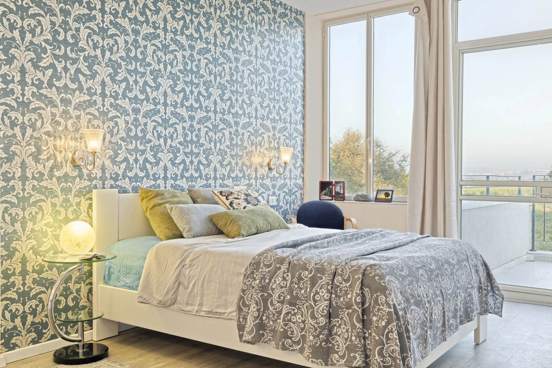 A bedroom features a white bed set against blue and white damask wallpaper, a glass bedside table, and large windows.