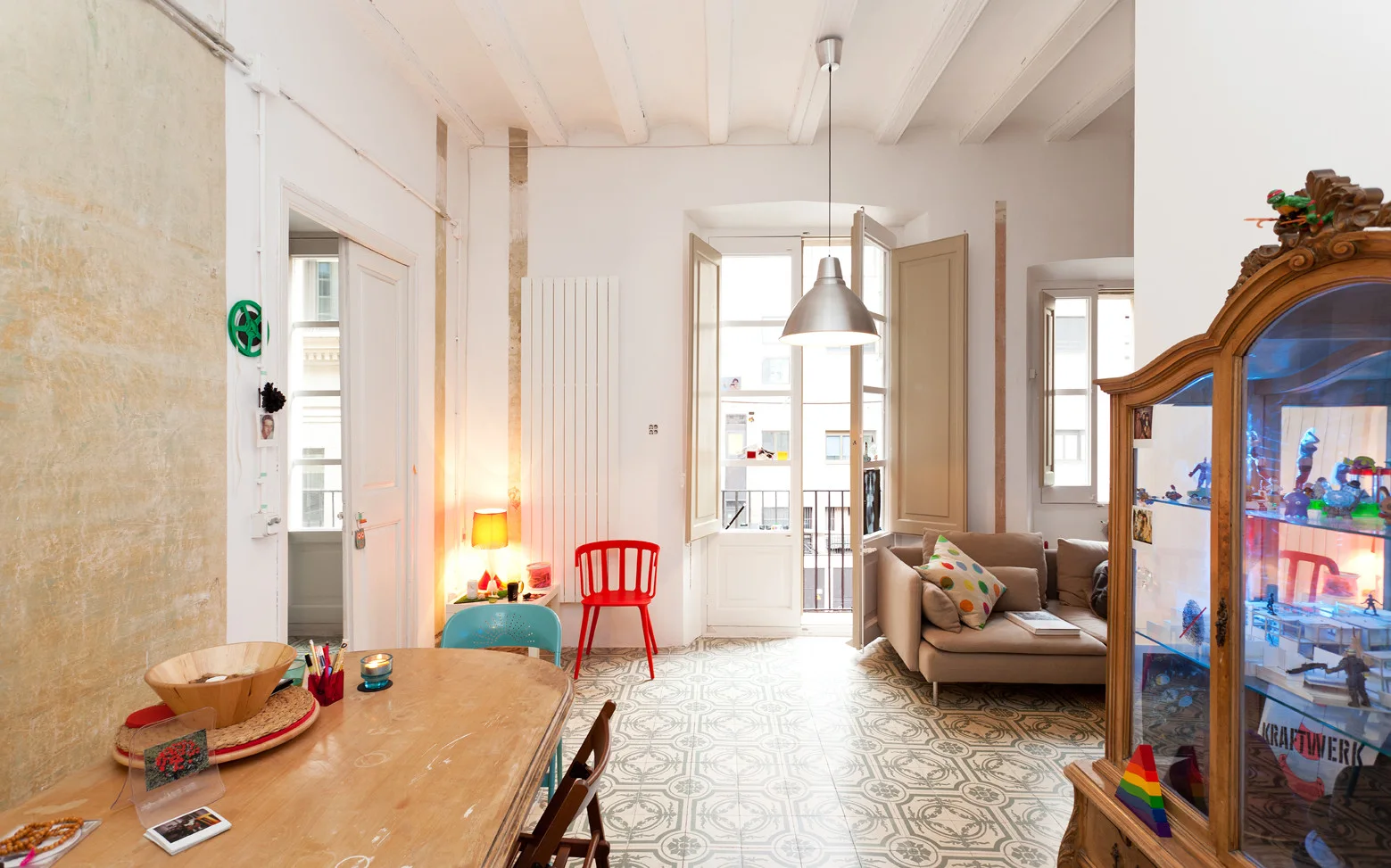 An eclectic apartment room featuring patterned floor tiles, white painted ceiling beams, a wooden dining table, and a beige sofa near a balcony window.
