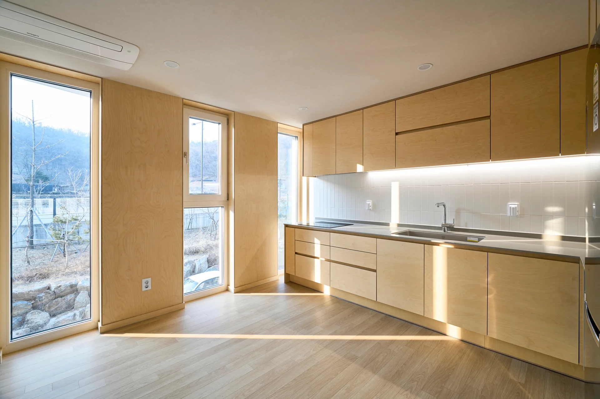 A minimalist kitchen features flat-panel light wood cabinetry, matching wood wall paneling, a white tiled backsplash, and large glass doors.