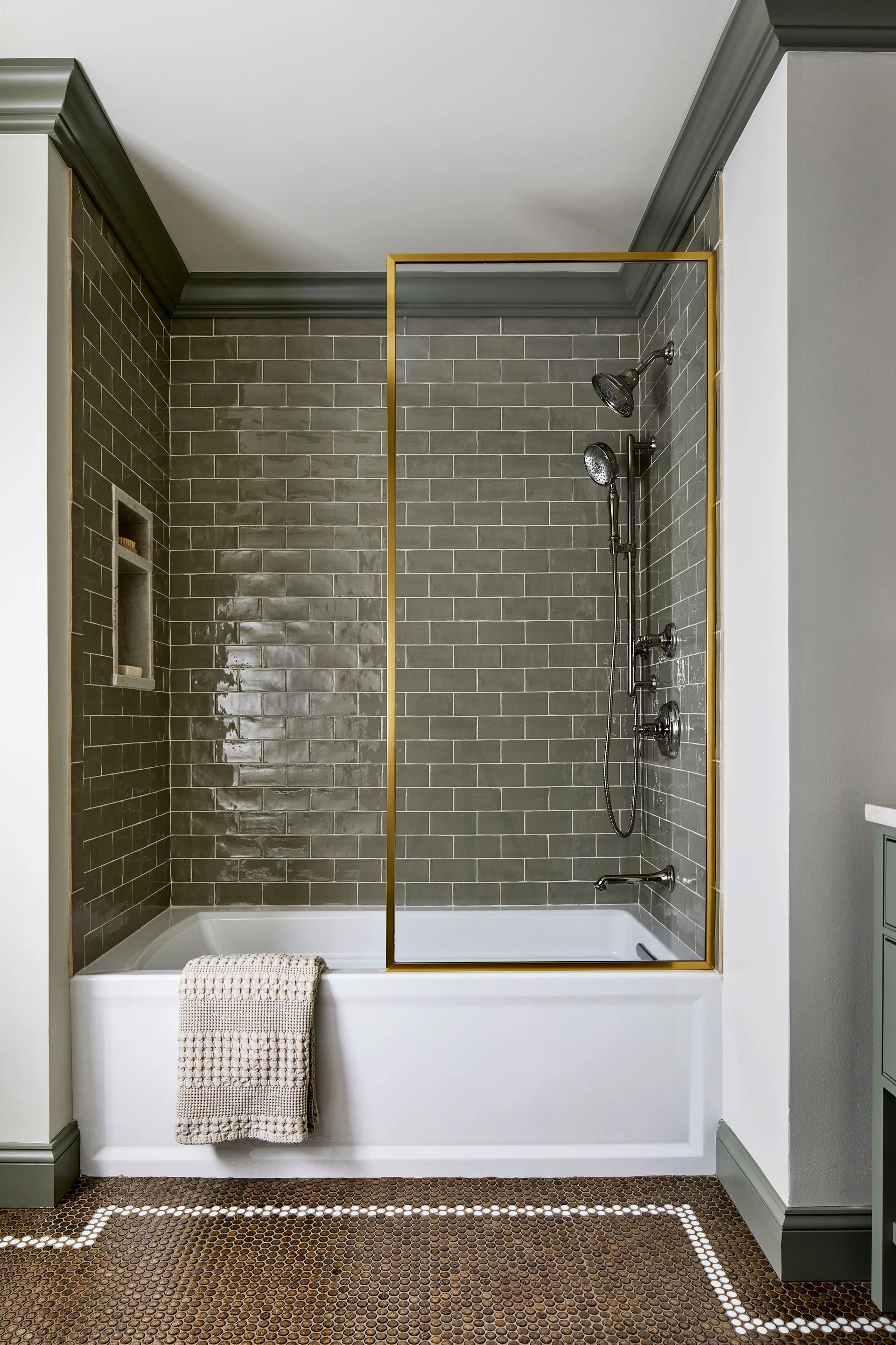 A bathroom featuring green glossy subway tiles on the walls, a white bathtub with a brass-framed glass partition, and brown penny tile flooring.