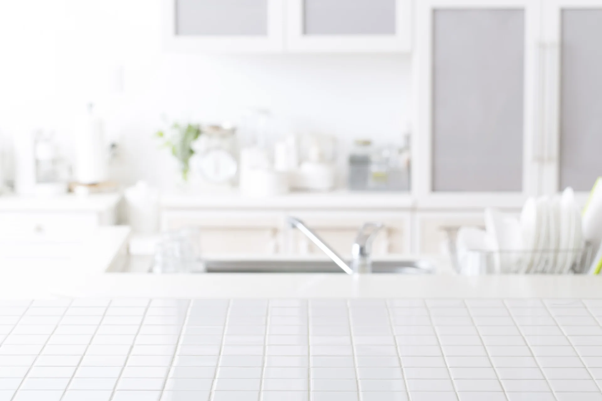 A close-up view of a bright white tiled kitchen countertop with a grid pattern, with a blurred sink faucet and white cabinets in the background.