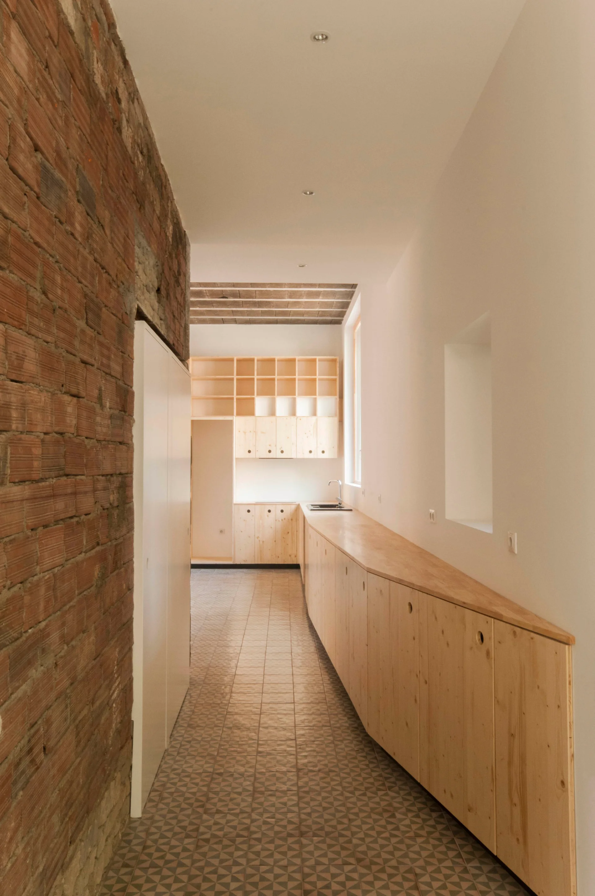 A narrow corridor kitchen featuring an exposed brick wall on one side, geometric tile flooring, and long light wood cabinets extending toward a sink.