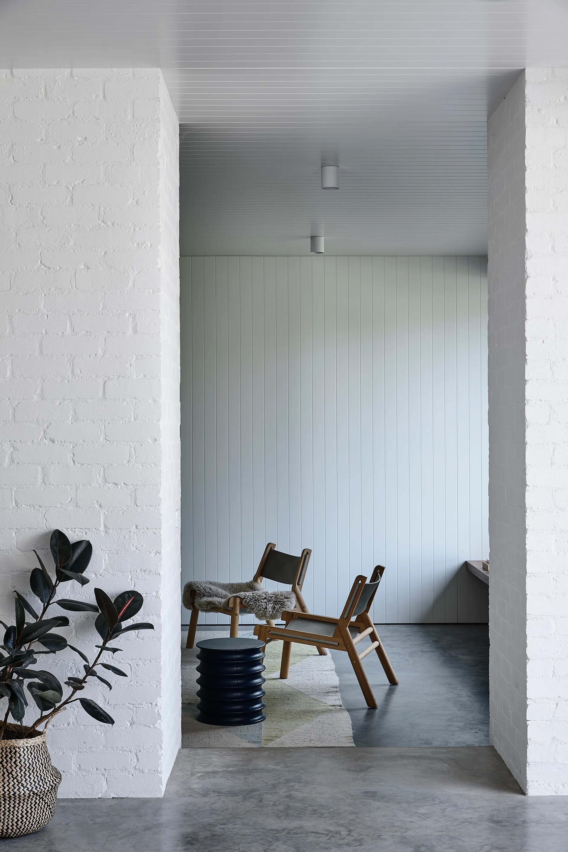 Wooden lounge chairs sit on a geometric rug in a minimalist space framed by white painted brick pillars and a vertical paneled wall.