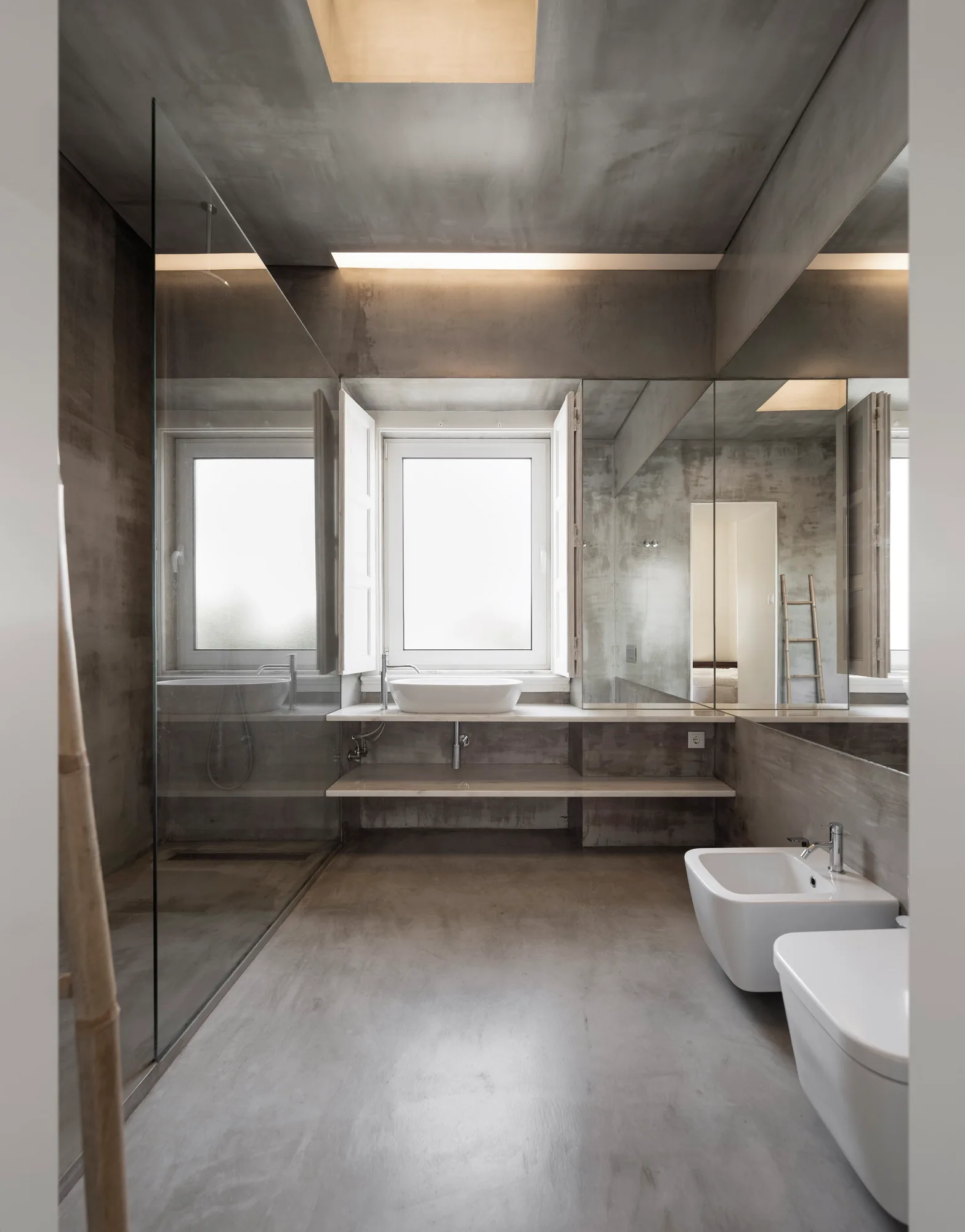 A grey concrete bathroom featuring a glass shower screen, a floating vanity with a white sink, and white ceramic fixtures.