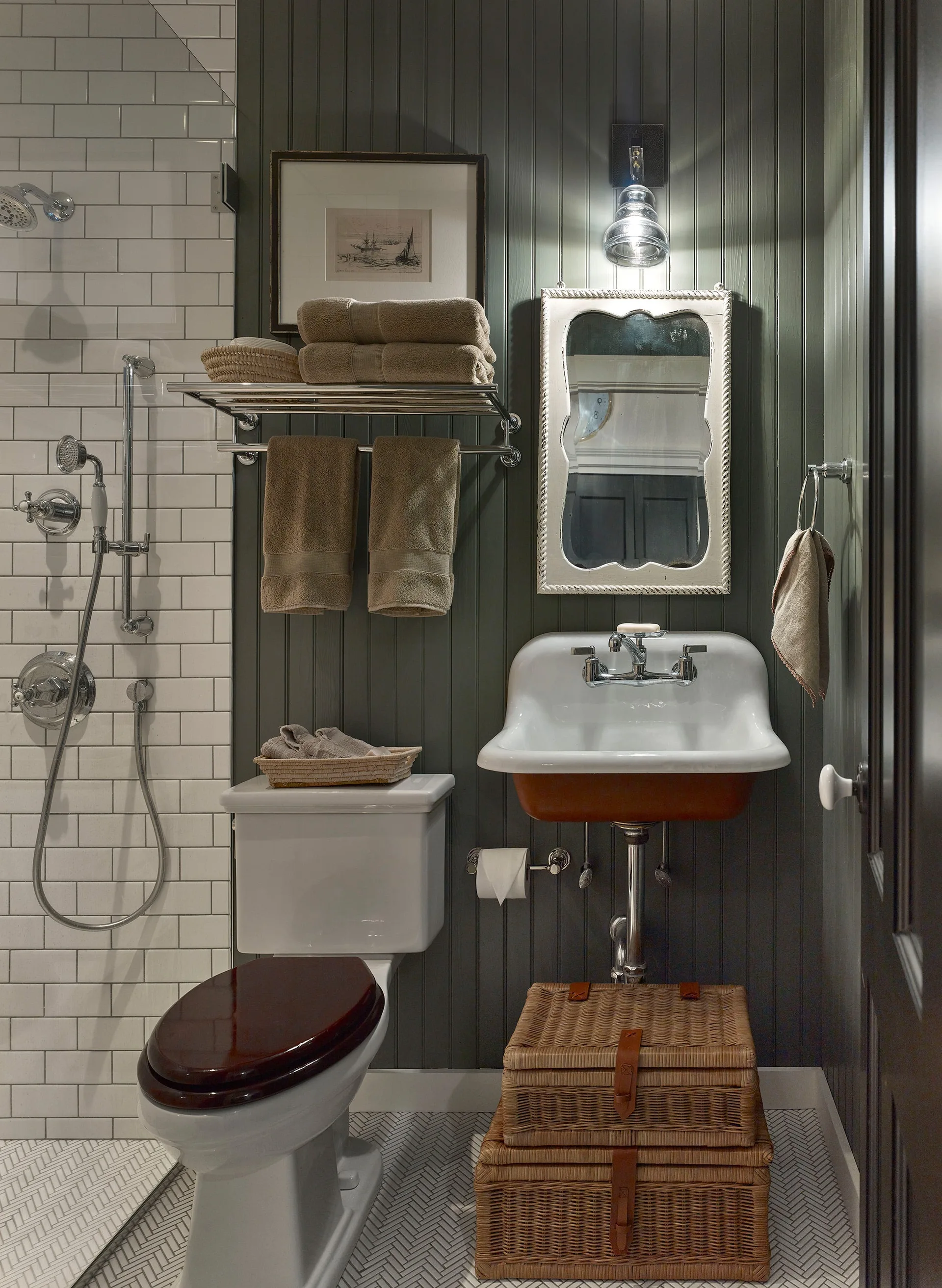 A small bathroom features dark green beadboard walls, a white wall-mounted sink, stacked wicker baskets, and a toilet with a dark wooden lid.