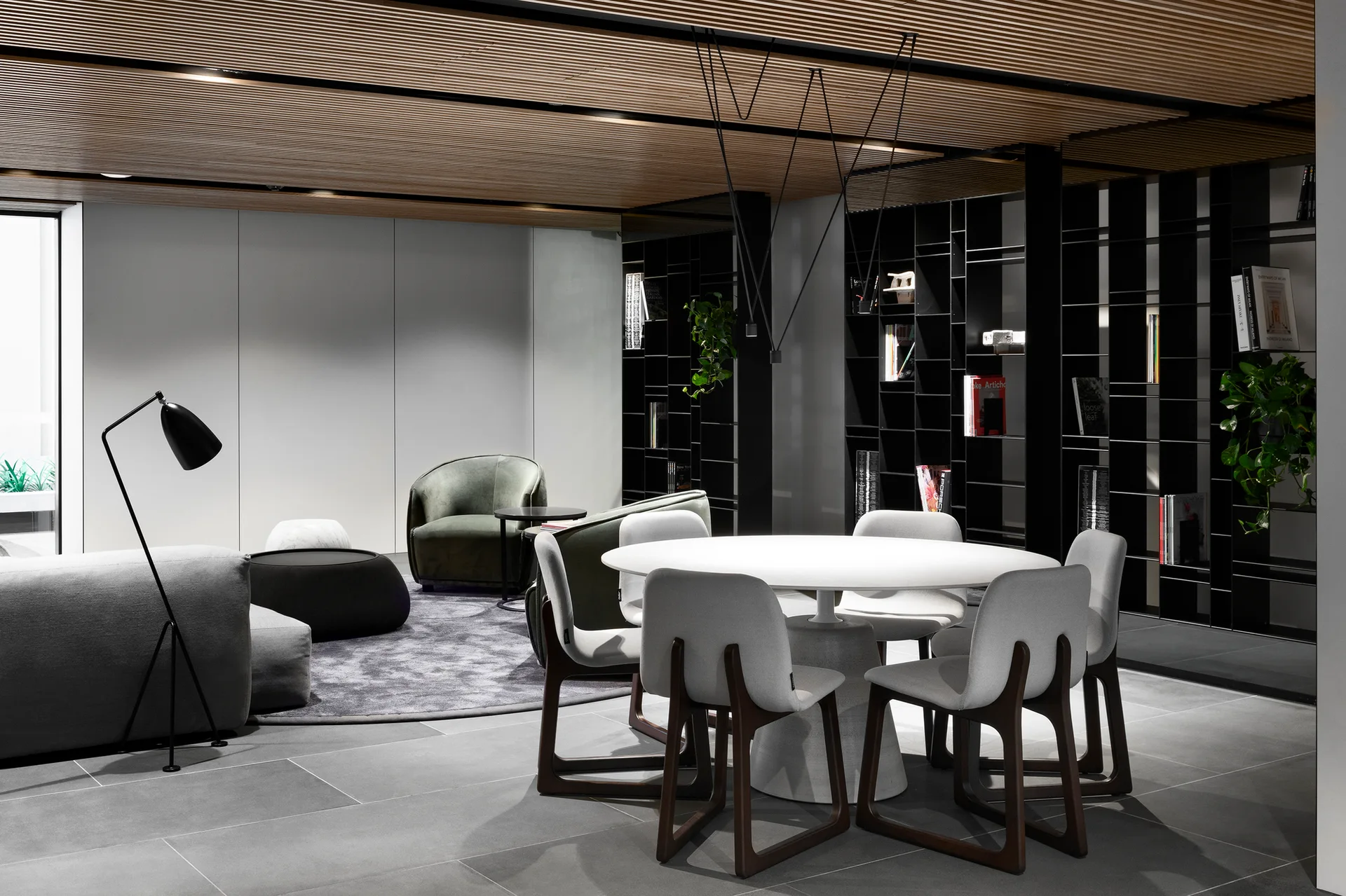 An open-plan dining room features a round white table, grey chairs, a black grid bookshelf, and a linear wood slatted ceiling.