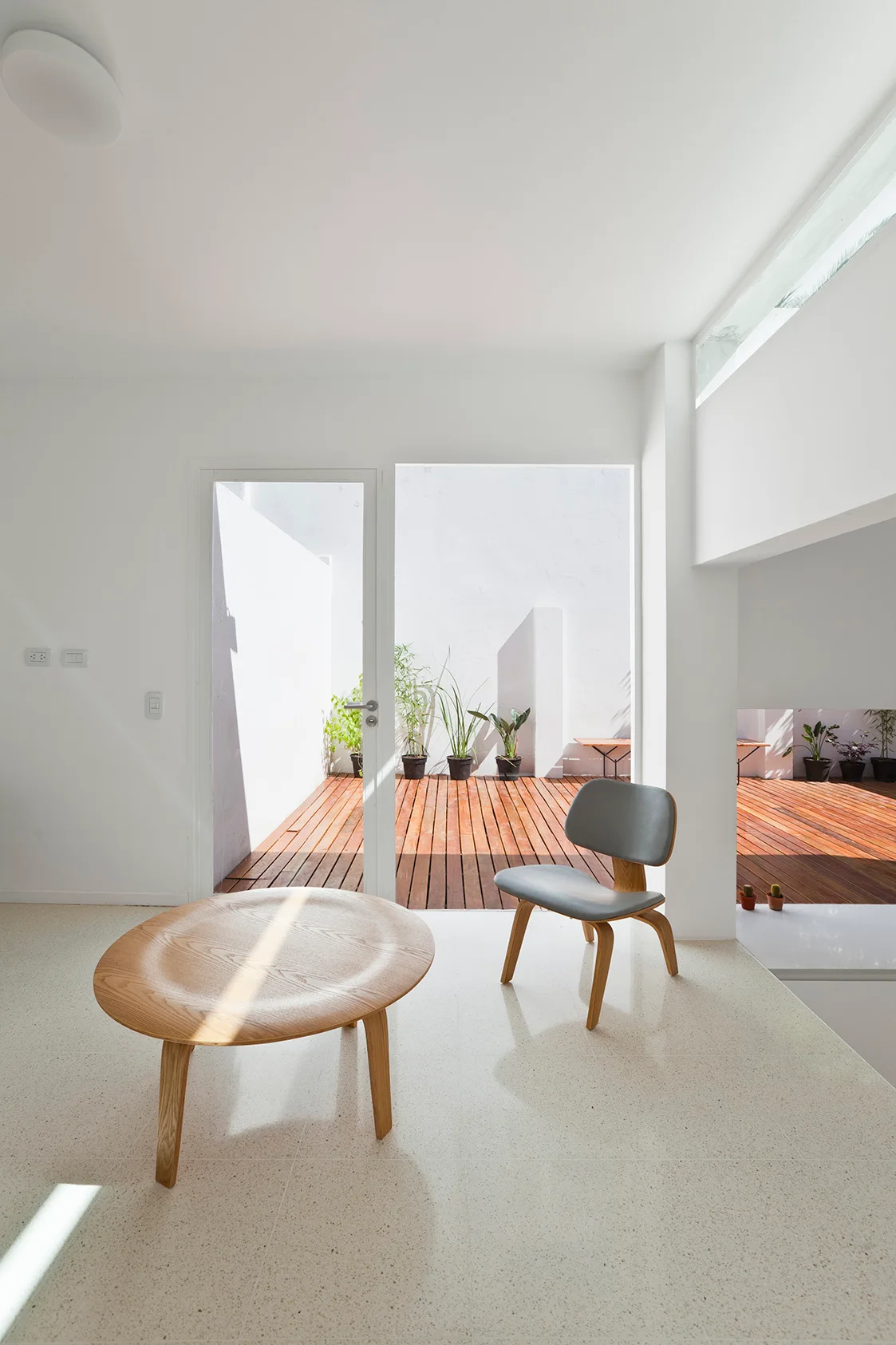 A minimalist white living room features a round wooden coffee table and grey chair facing a glass door that opens onto a sunny wooden deck with plants.