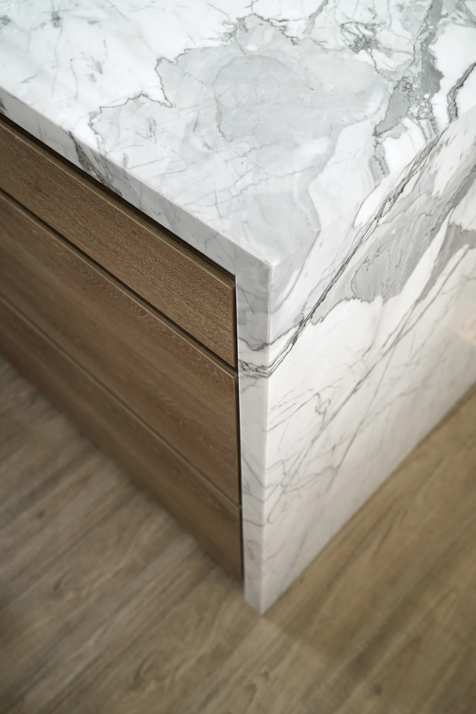 A close-up view of a kitchen island featuring a white marble waterfall countertop next to warm wood cabinet drawers.