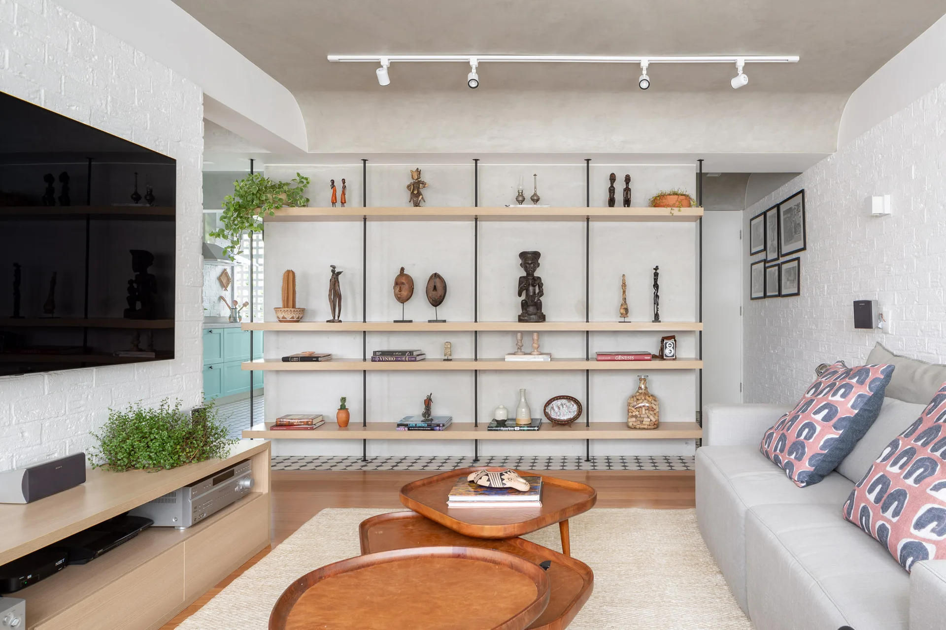 A light grey sofa with patterned pink pillows faces nested wood coffee tables in a modern living room featuring open shelving and a white brick wall.