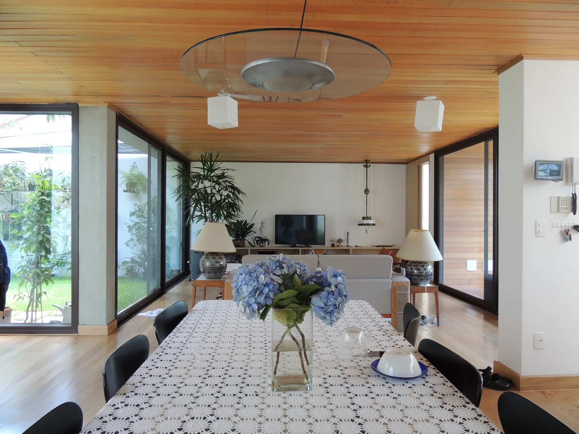 A modern dining room features a lace-covered table with blue flowers, black chairs, and a slatted wood ceiling.