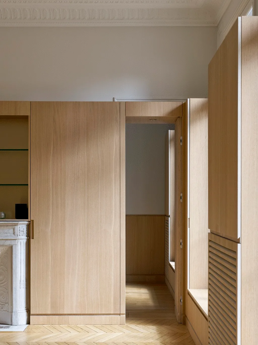 A light oak-paneled wall with built-in storage cabinets, a transition doorway, herringbone flooring, and detailed white ceiling trim.