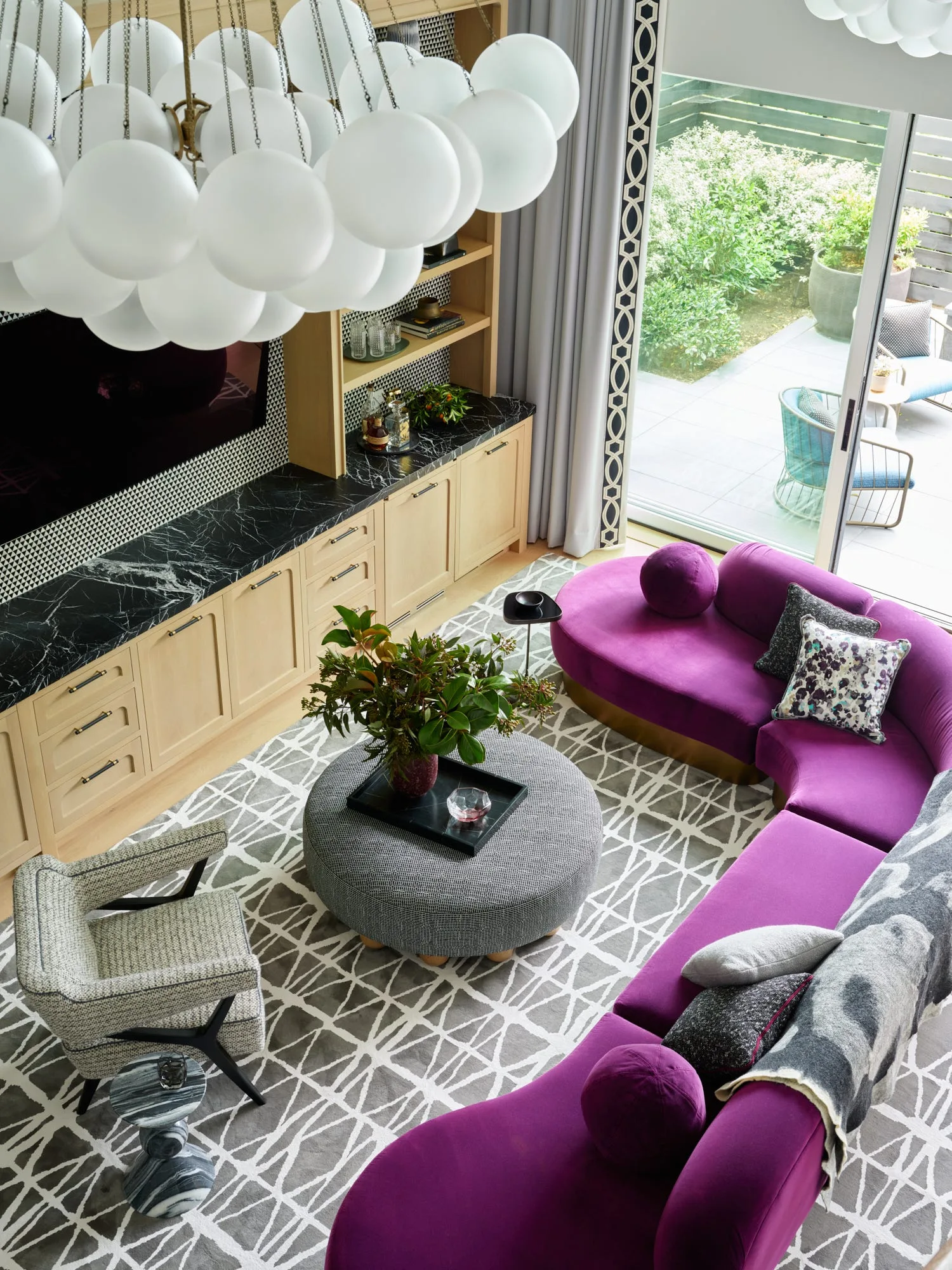 A high-angle view of a living room with a curved purple sofa, a round grey patterned ottoman, and custom light wood cabinetry with black marble.