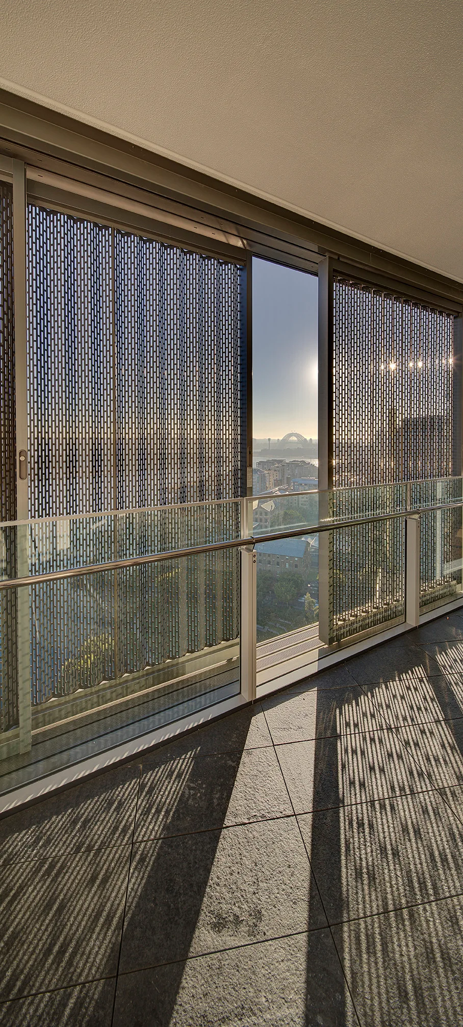 A sunlit apartment balcony featuring dark tiled floors, a glass balustrade, and sliding metal mesh privacy screens that cast striped shadows.