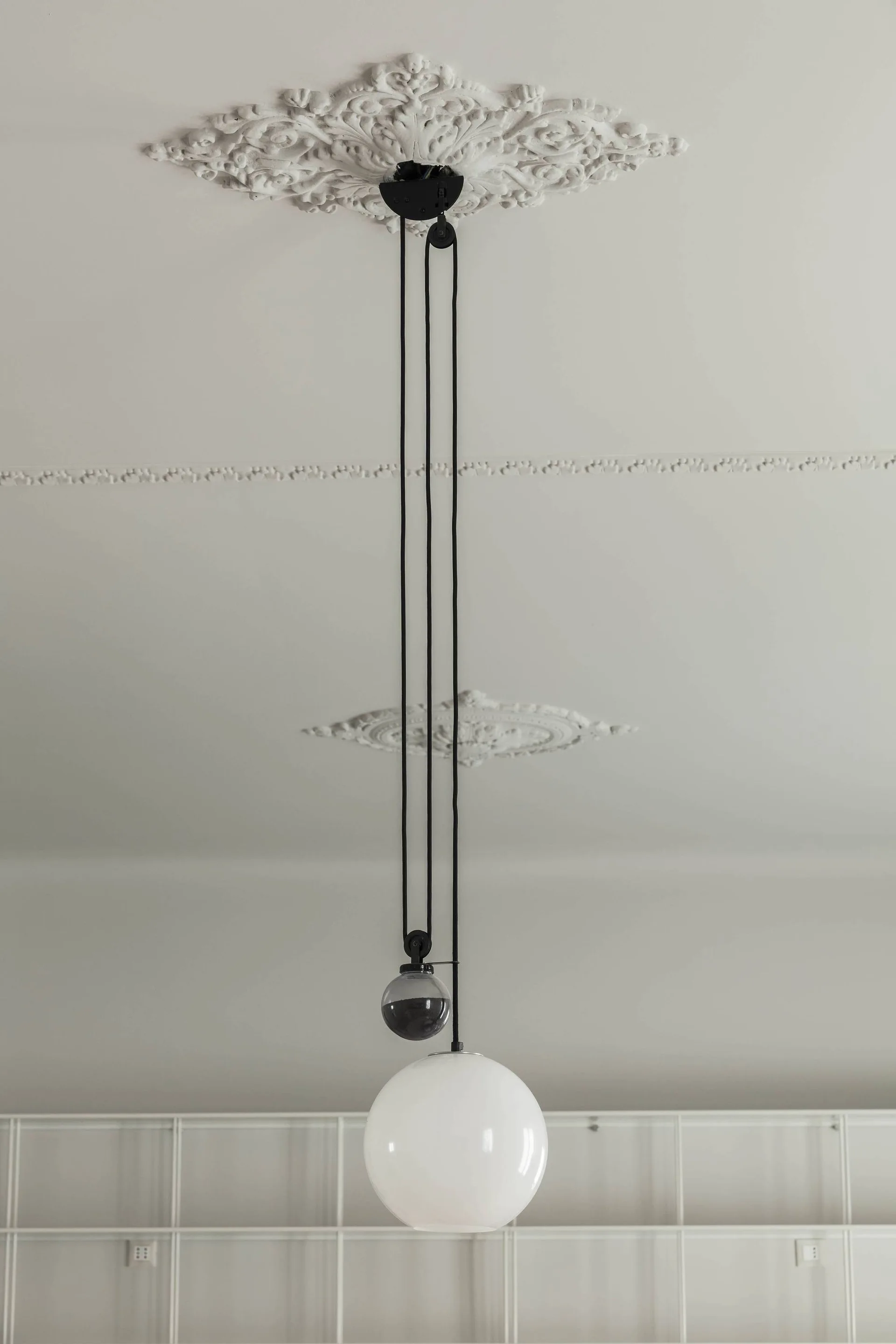 A modern black pulley pendant light with a white globe hangs from an ornate white plaster ceiling medallion.