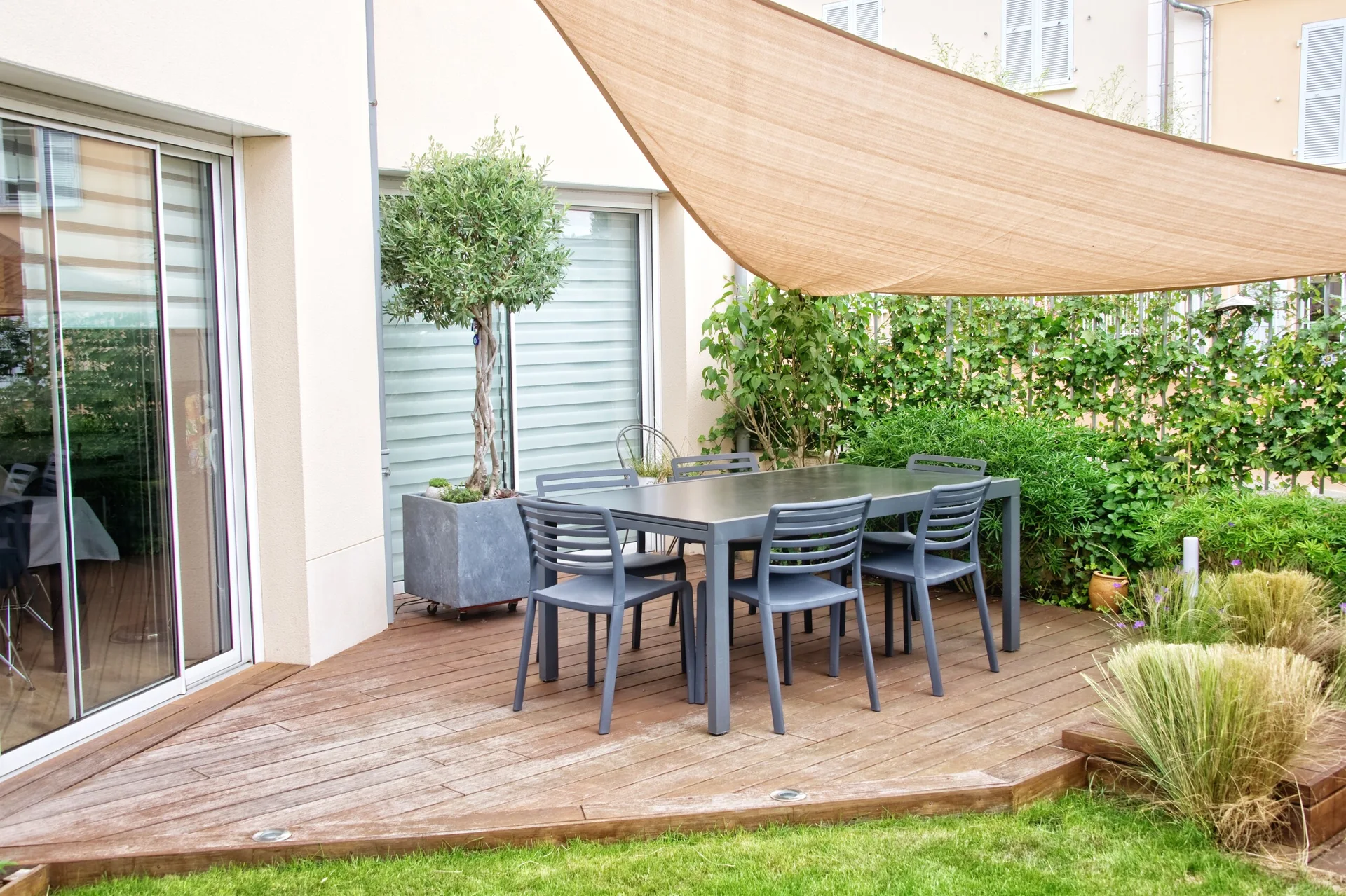 A dark grey metal dining set sits on a wooden deck beneath a large beige triangular sun shade sail canopy next to a green lawn.