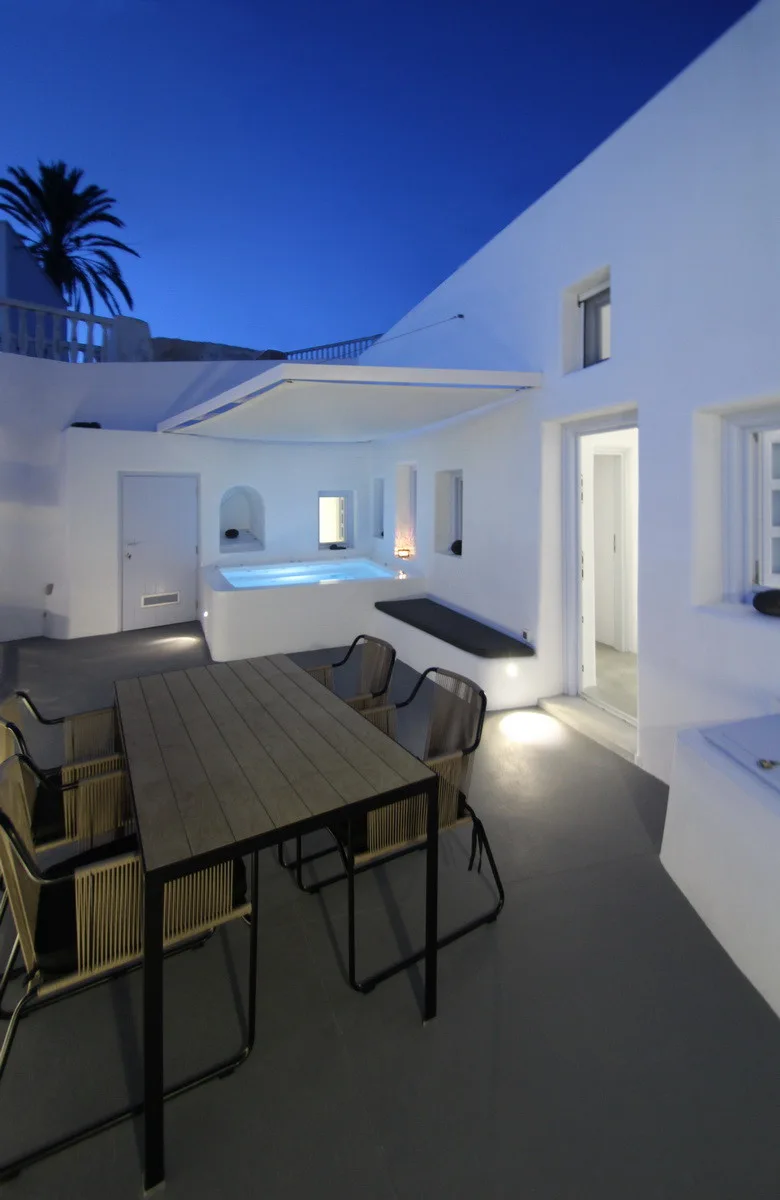 A white stucco patio at twilight featuring a wooden dining table, modern chairs, a built-in bench, and an illuminated plunge pool.