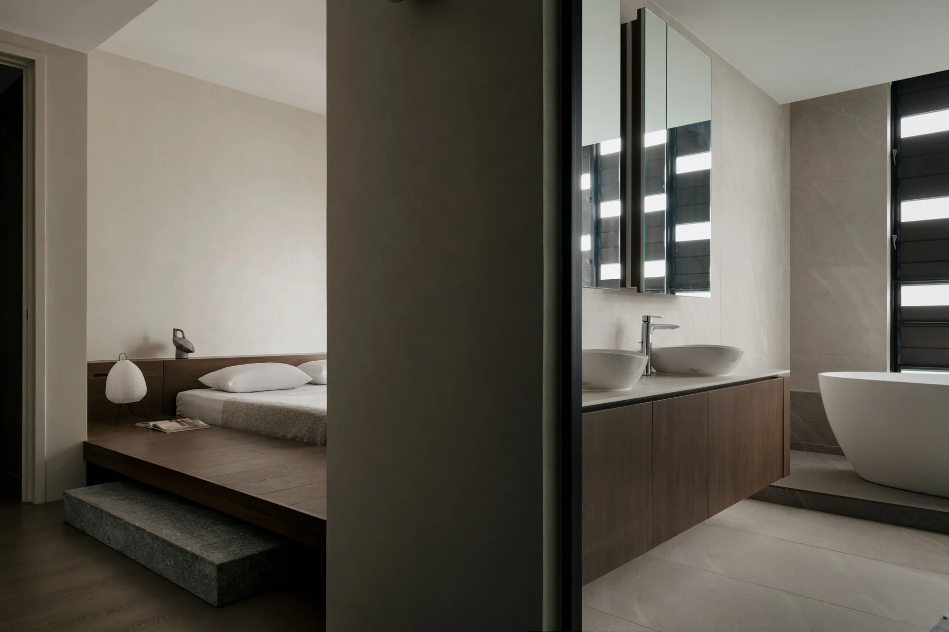 A minimalist bedroom with a low wood platform bed next to a contemporary bathroom featuring dual vessel sinks and a freestanding tub.