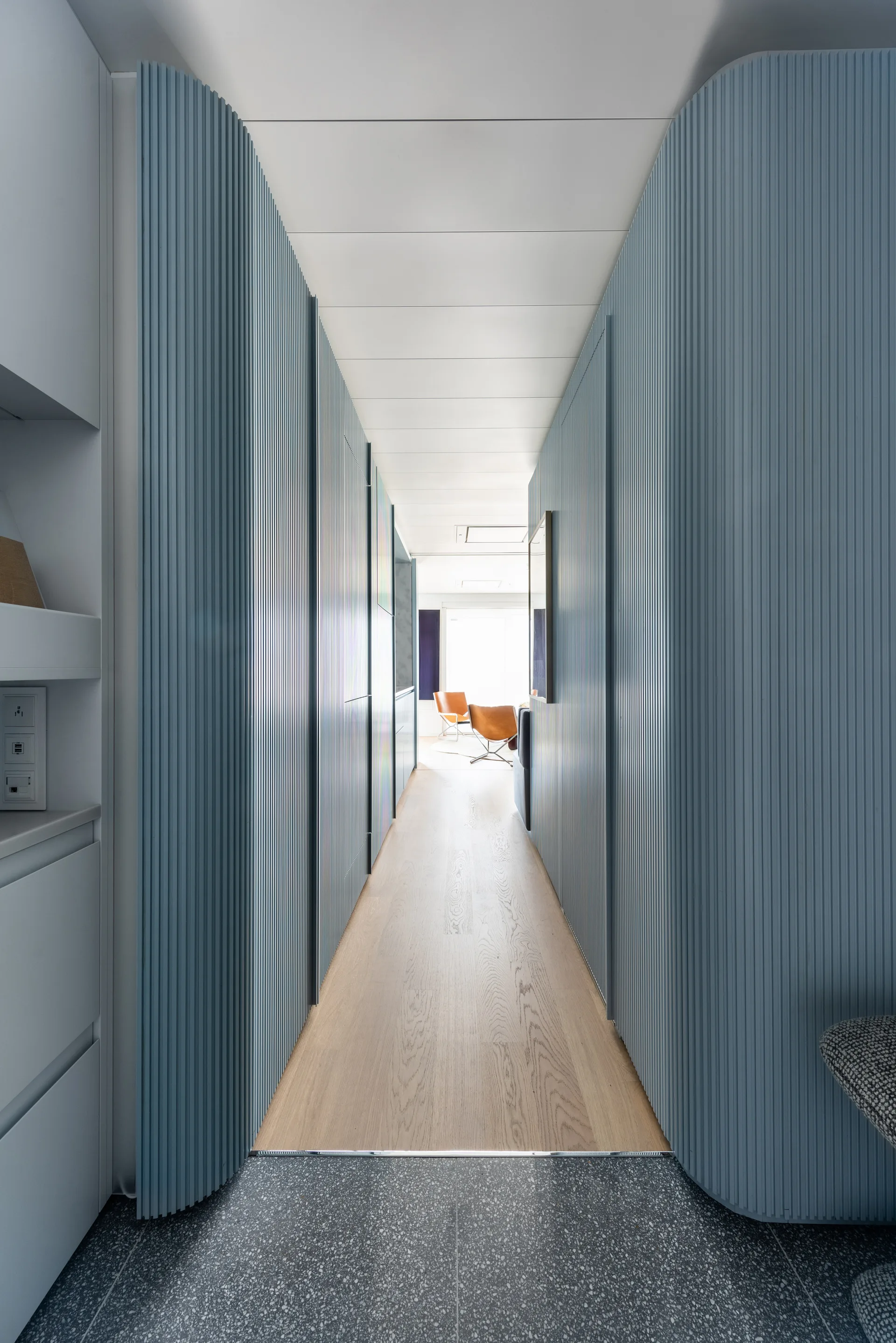 A narrow hallway features curved blue fluted wall panels, light wood flooring, and a terrazzo threshold looking into a sunlit room.