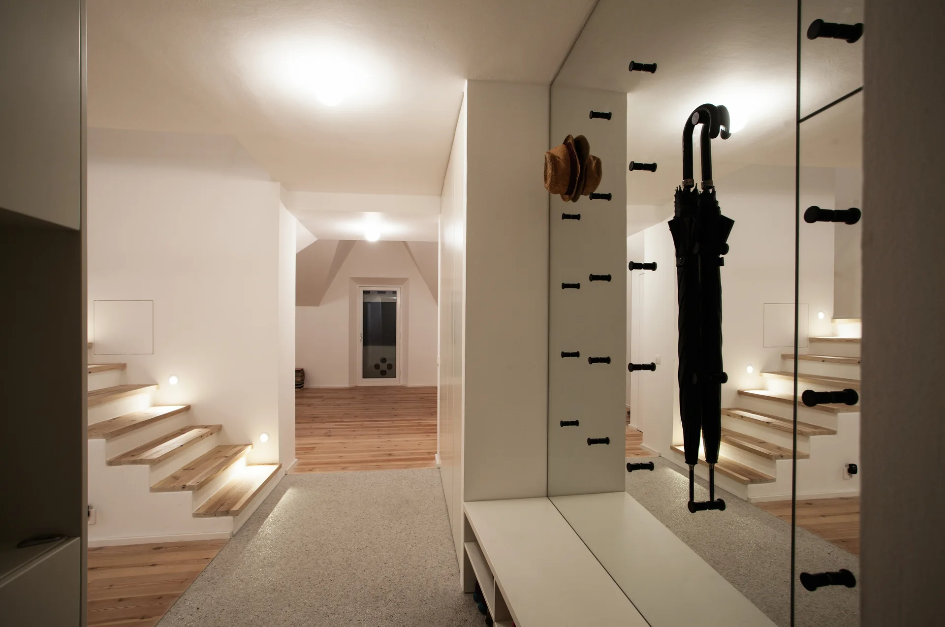 A minimalist white entryway features a large wall mirror with black pegs, a white built-in bench, and wooden stairs with warm recessed lighting.