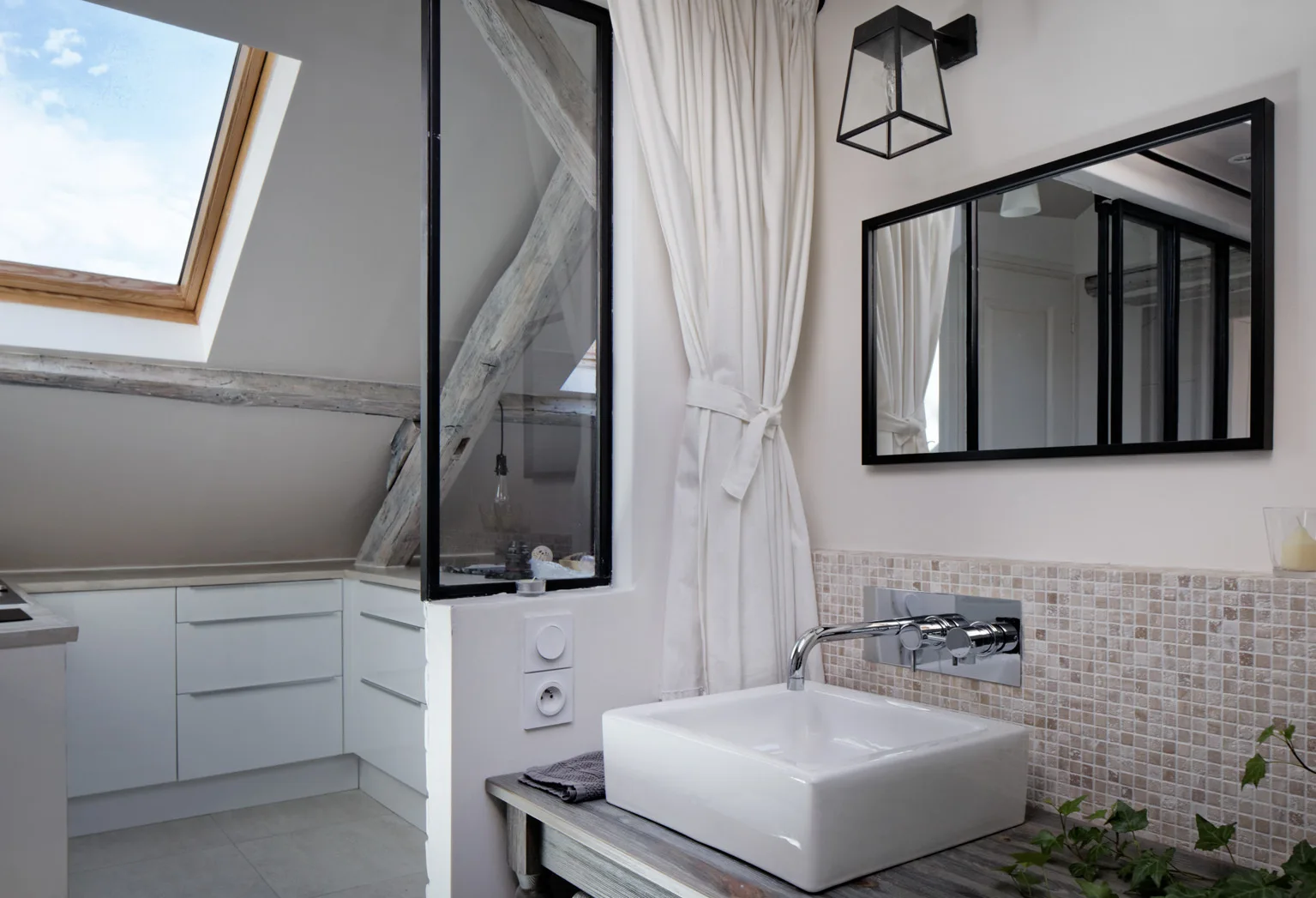 A bathroom vanity features a square vessel sink, beige mosaic tile backsplash, black-framed mirror, and a sloped ceiling with a skylight.