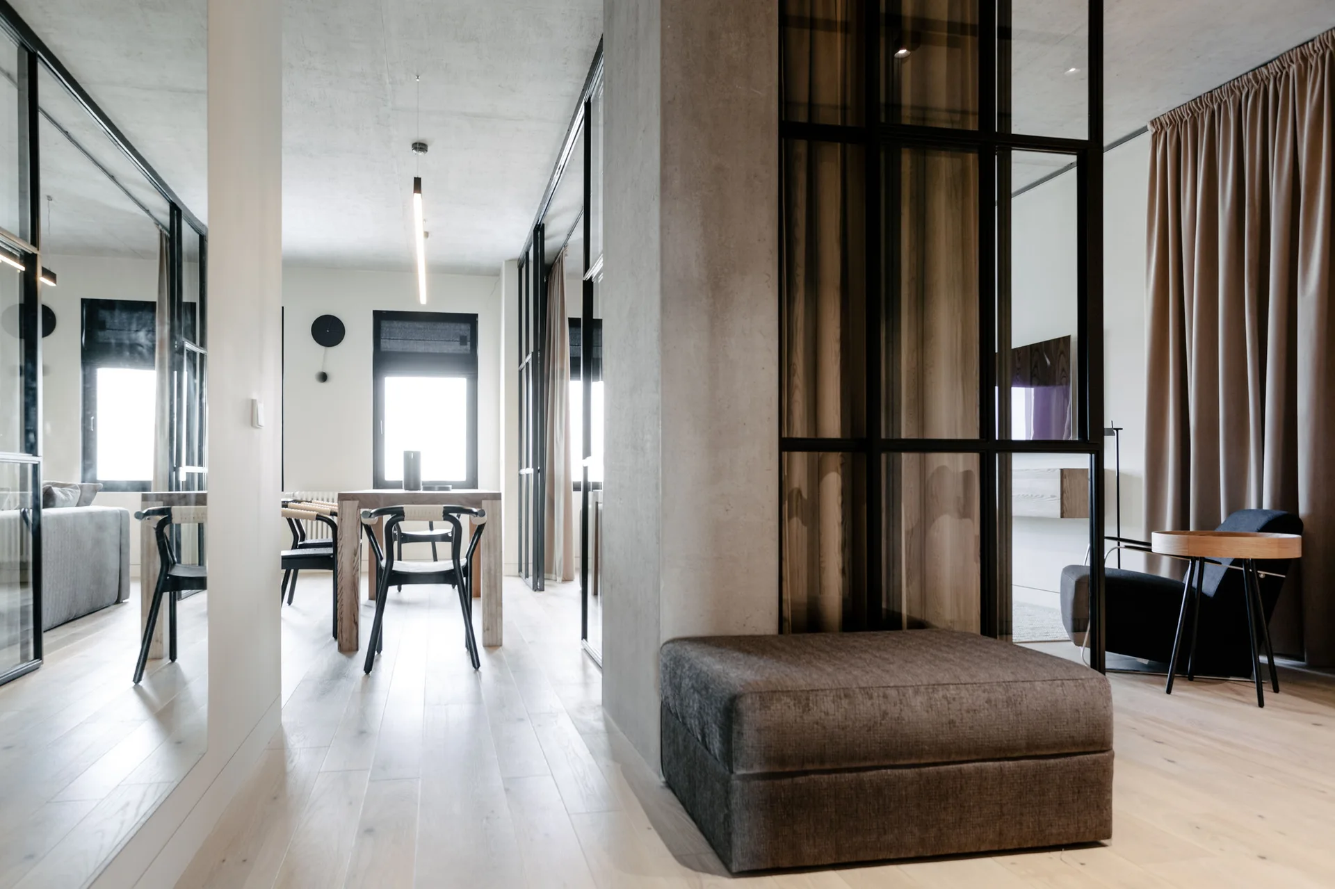 An open-concept residential interior with light wood flooring, a central concrete column, a grey ottoman, and black-framed glass partition walls.