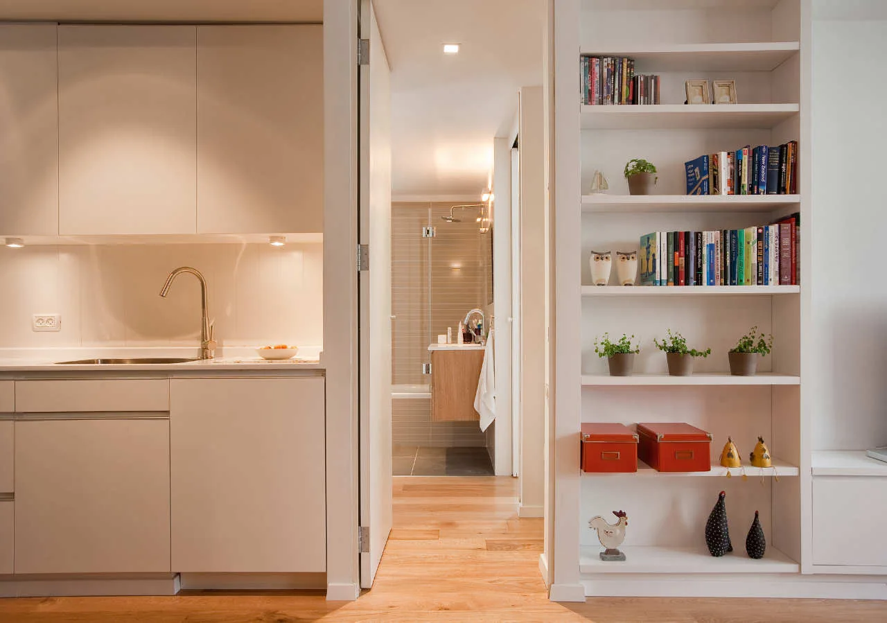 White flat-panel kitchen cabinets with a gooseneck faucet stand next to a built-in bookshelf and an open doorway leading to a bathroom.