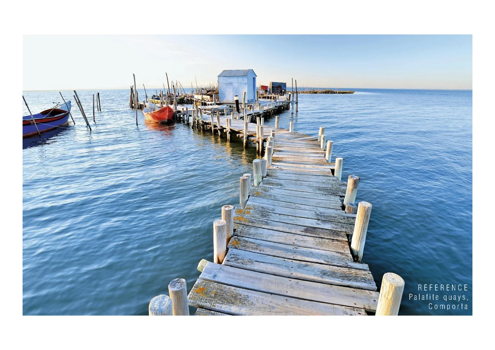 A weathered wooden dock with vertical timber posts extends into calm blue water with small boats and a tiny light blue shed.