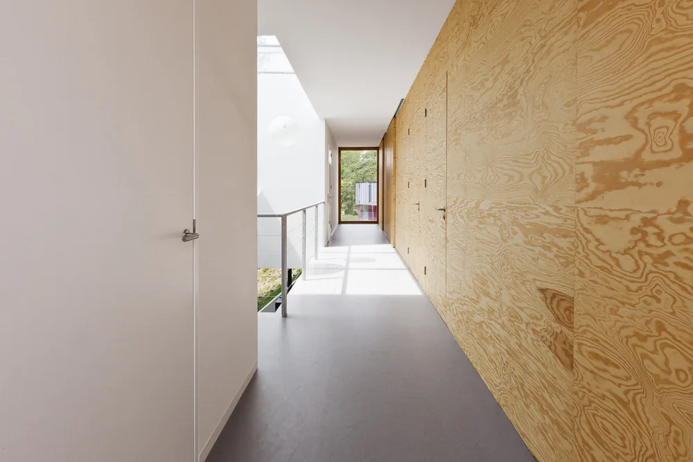 A minimalist hallway with light grey flooring, white walls on one side, and a continuous wall of plywood panels with integrated flush doors.