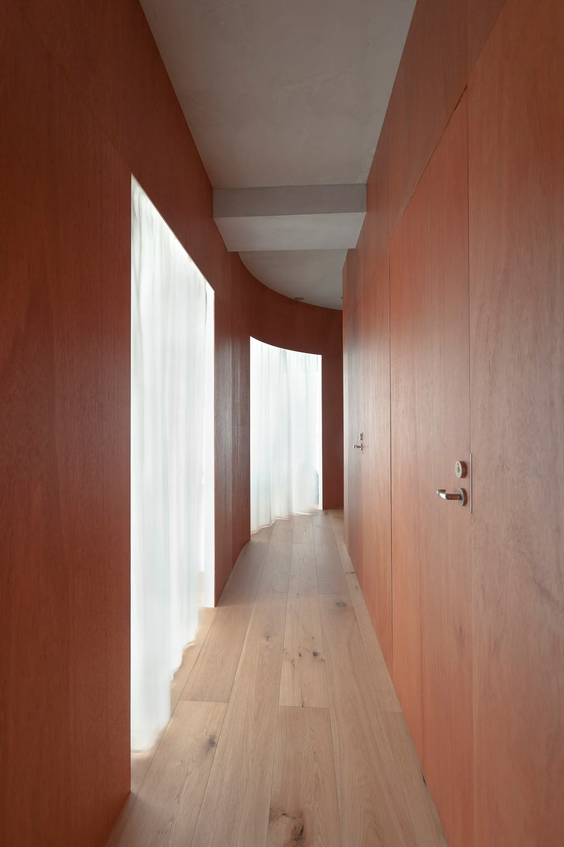 A curved hallway features floor-to-ceiling warm wood wall paneling, light wood flooring, flush doors, and inset sheer white curtains filtering natural light.