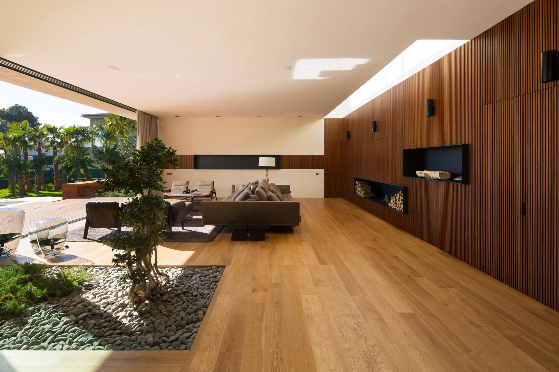 A spacious minimalist living room featuring a vertical wood slat accent wall, hardwood floors, a grey sofa, and an indoor pebble garden with a small tree.