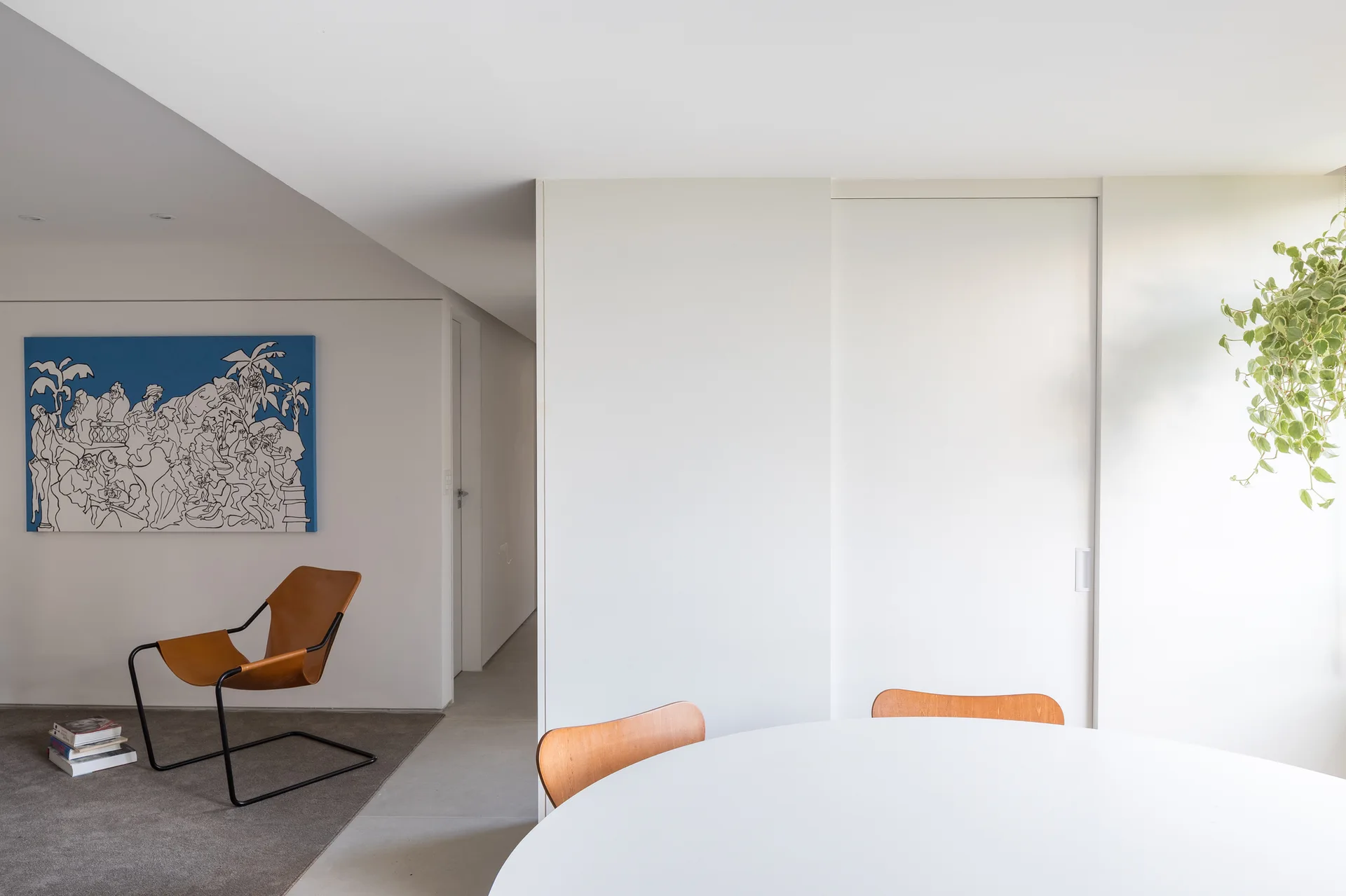 A minimalist open-plan dining space with a white round table, wooden chairs, a tan leather armchair on a gray rug, and a large blue painting on the wall.