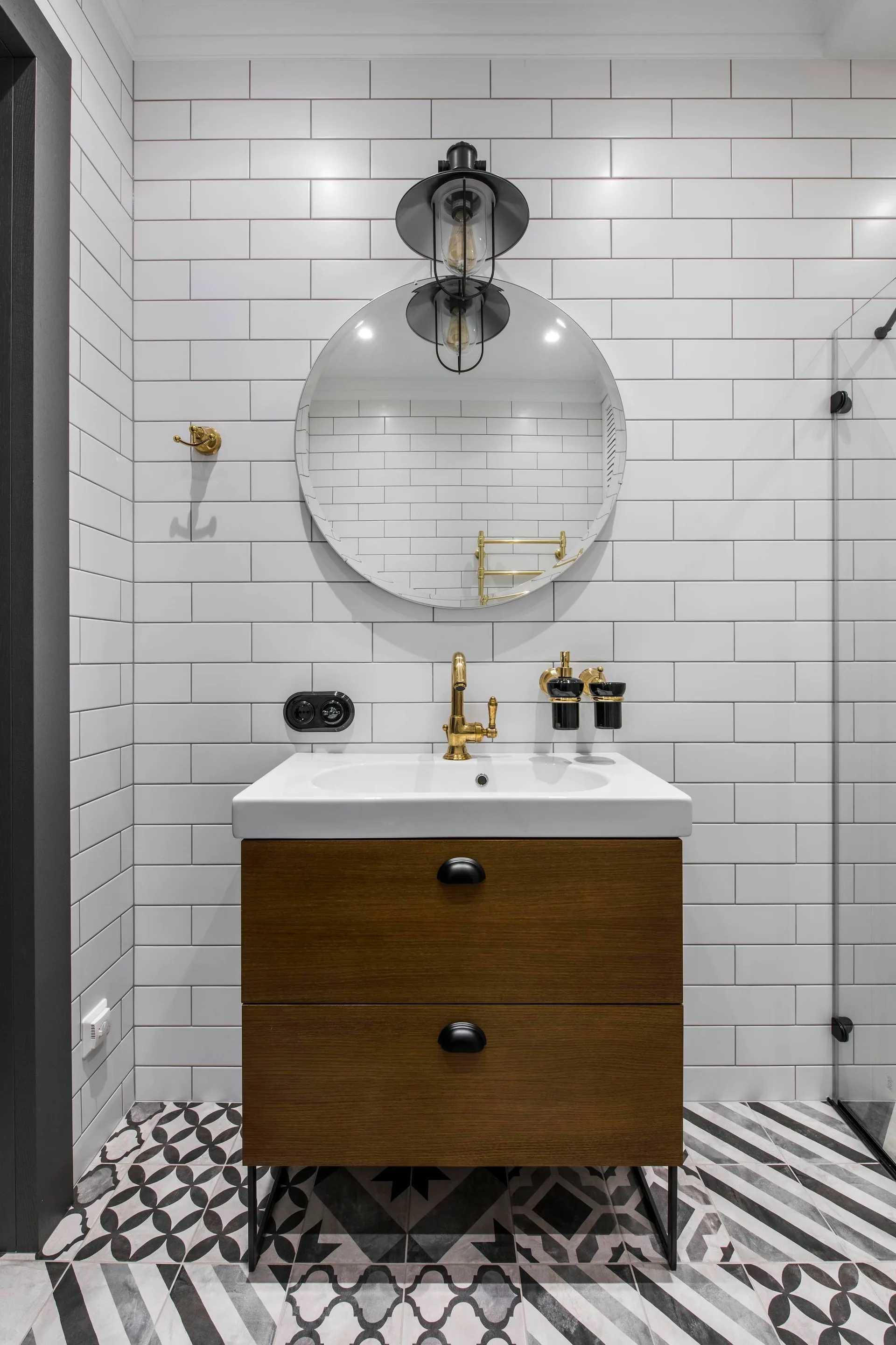 A wood bathroom vanity with black handles sits against a white subway tile wall under a round mirror and black industrial sconce.