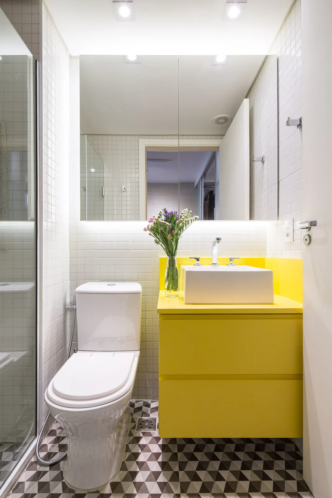A modern bathroom showing a bright yellow floating vanity, a white vessel sink, a white tiled wall, and geometric patterned floor tiles.
