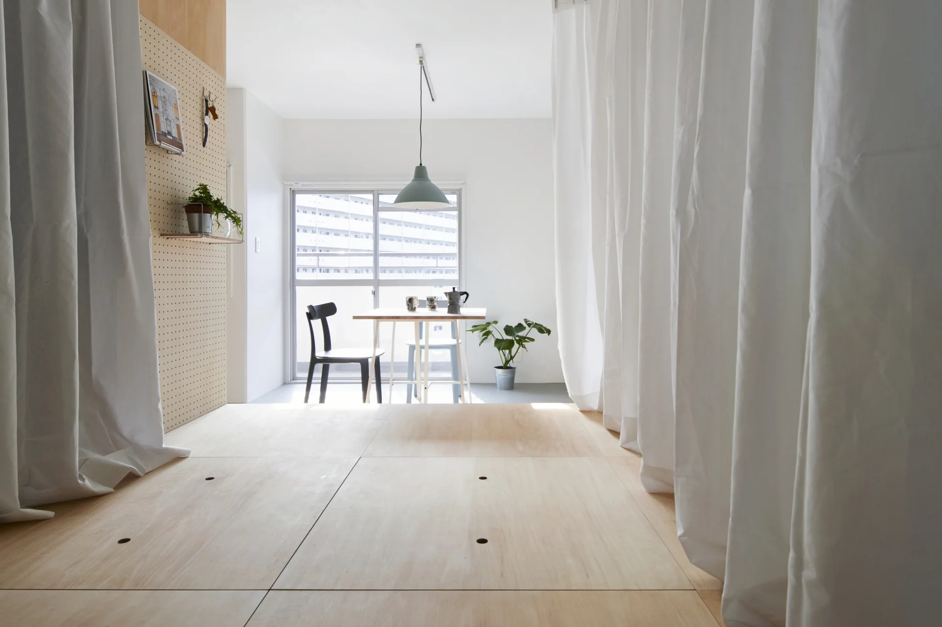 A minimalist dining area with a table and chairs sits beyond a raised plywood platform flanked by long white curtains.