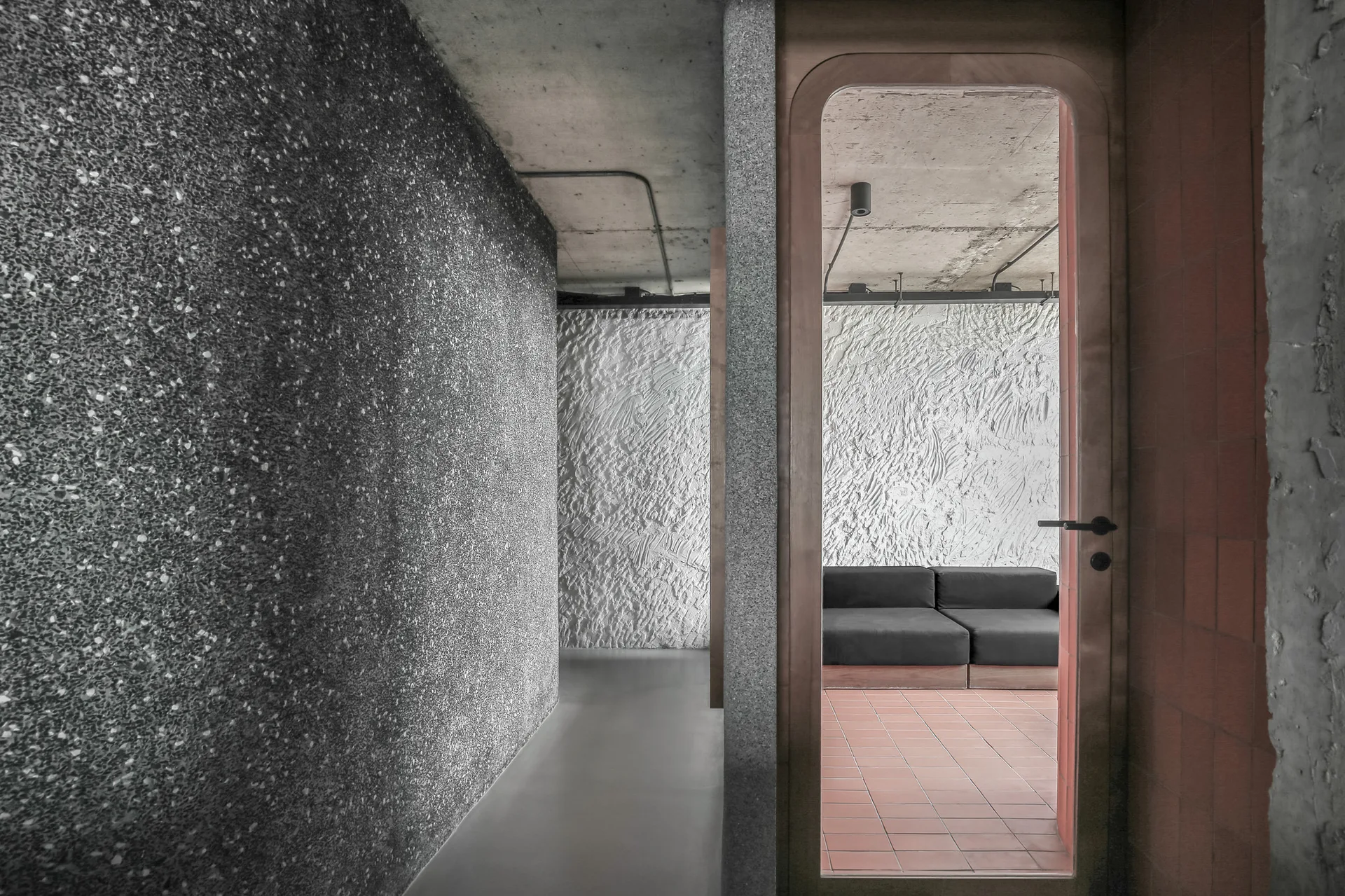 A view through a wood-framed doorway showing a dark sofa on terracotta tiles against a textured plaster wall, bordered by raw concrete and stone walls.